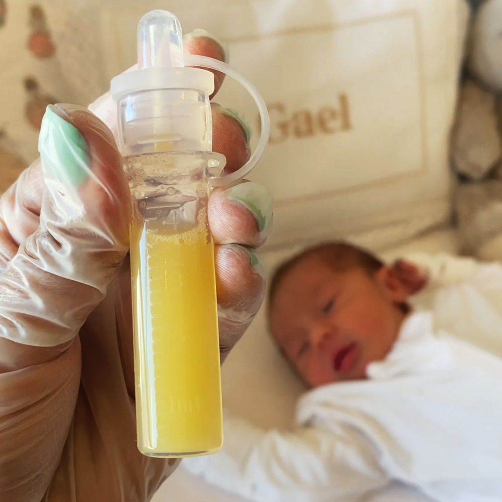 Eine Hand hält einen transparenten Behälter mit gelblicher Flüssigkeit. Ein schlafendes Baby im Hintergrund.