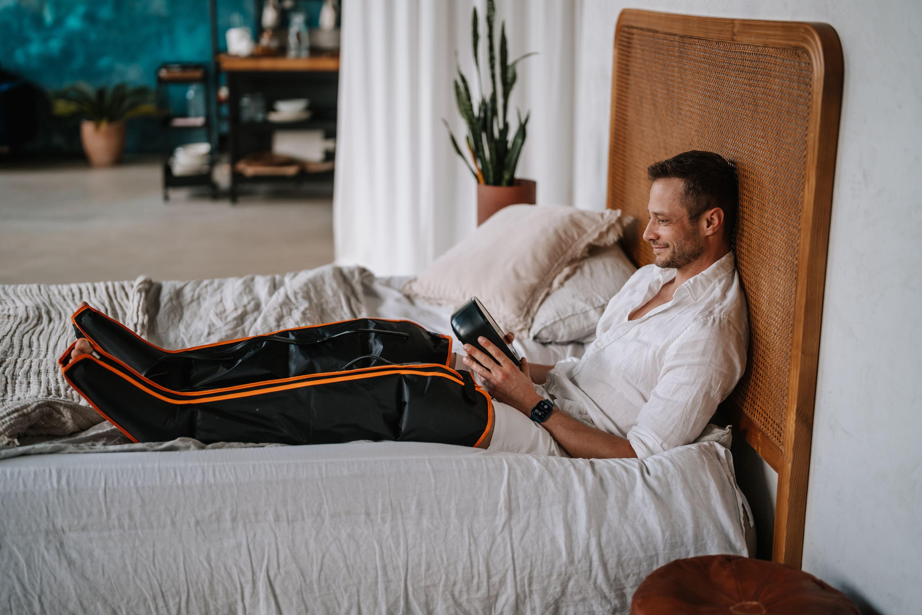 Mann liegt auf dem Bett mit Bein-Lymphmassagegerät. Schwarzes Gerät mit orangefarbenen Akzenten. Hält ein Tablet.