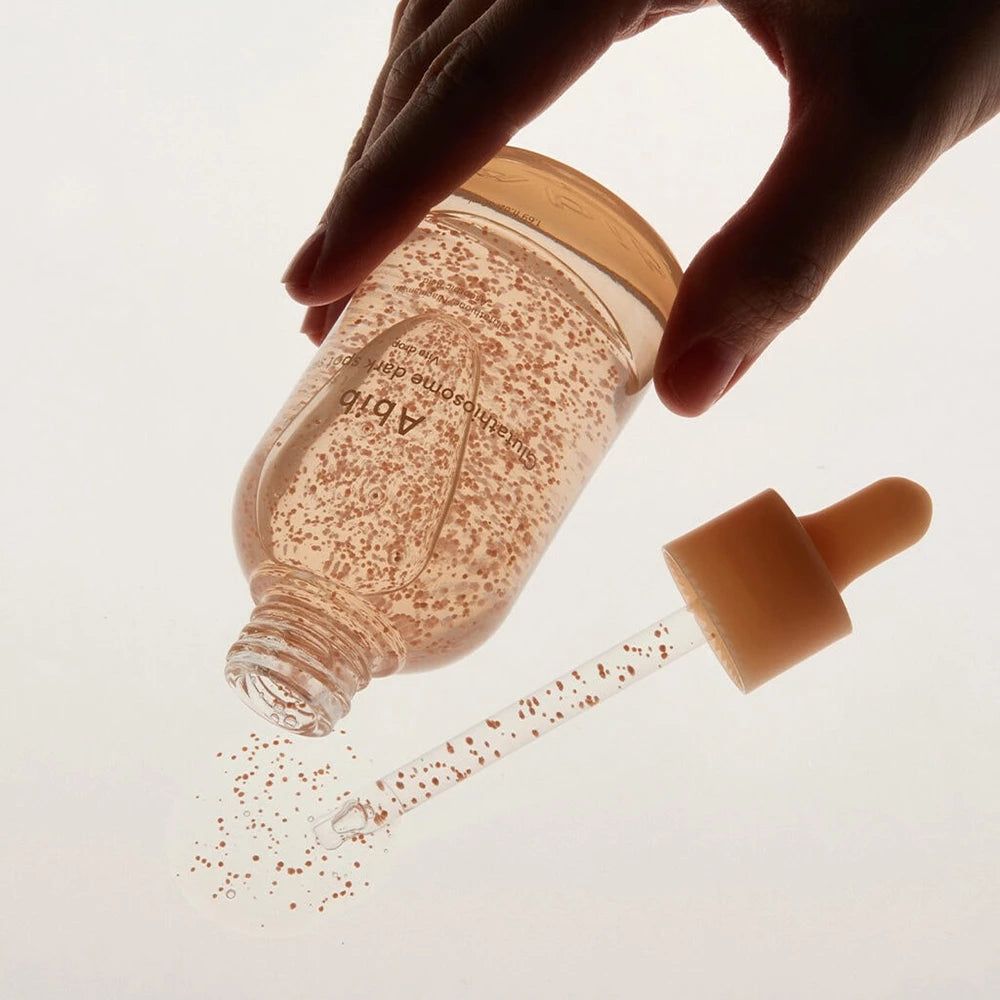 Glasflasche mit Serum, rosa Kügelchen, Pipette. Eine Hand hält die Flasche. Serum wird auf eine Oberfläche gegossen.