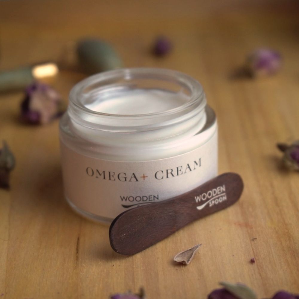 Glasbehälter mit Creme und Holzspatel. Aufschrift OMEGA+ CREAM und Wooden Spoon. Holzspatel neben dem Behälter.