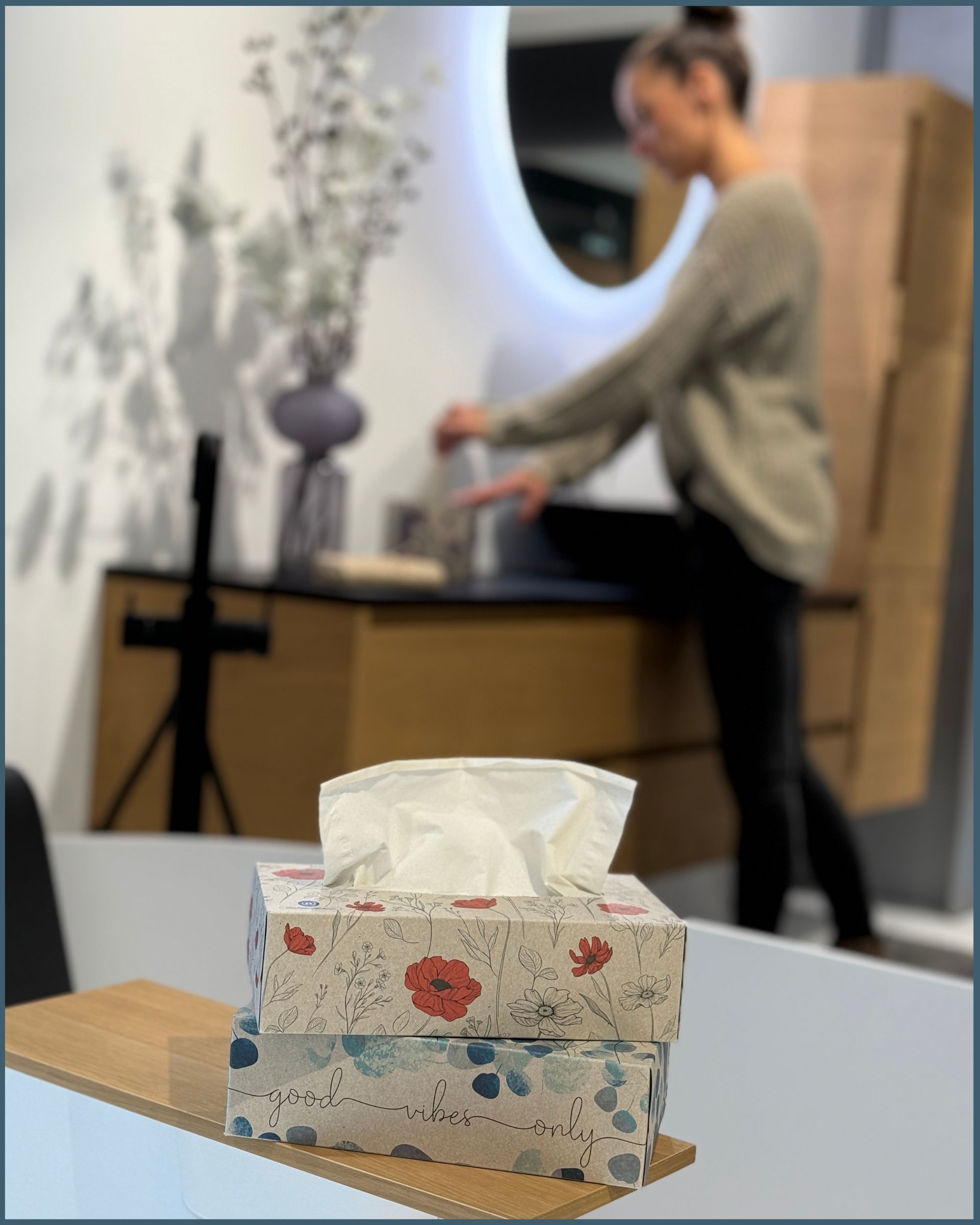 Zwei Packungen Taschentücher auf einer Holzablage. Eine Person im Hintergrund. Floral bedruckte Verpackung.