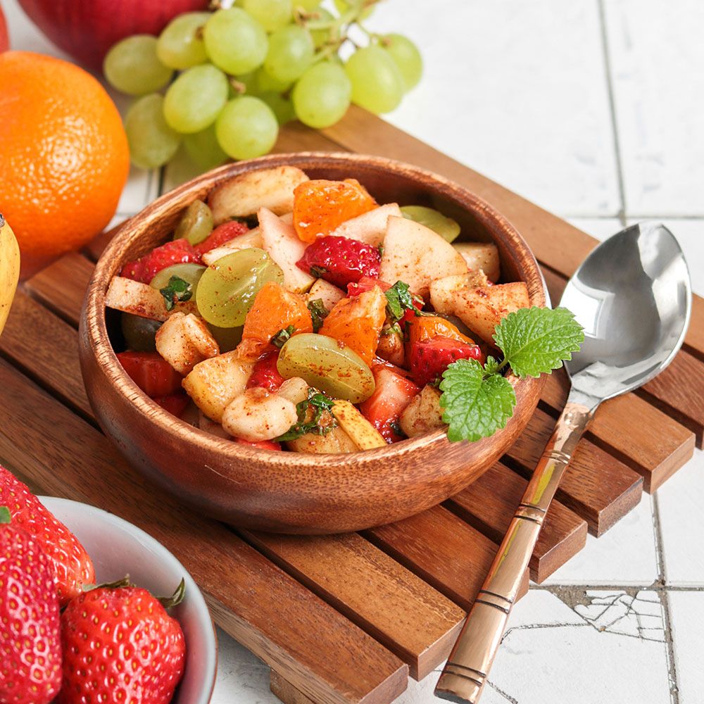 Obstsalat in Holzschale. Enthält Erdbeeren, Trauben, Orangen und Minze. Auf Holzbrett mit Löffel.