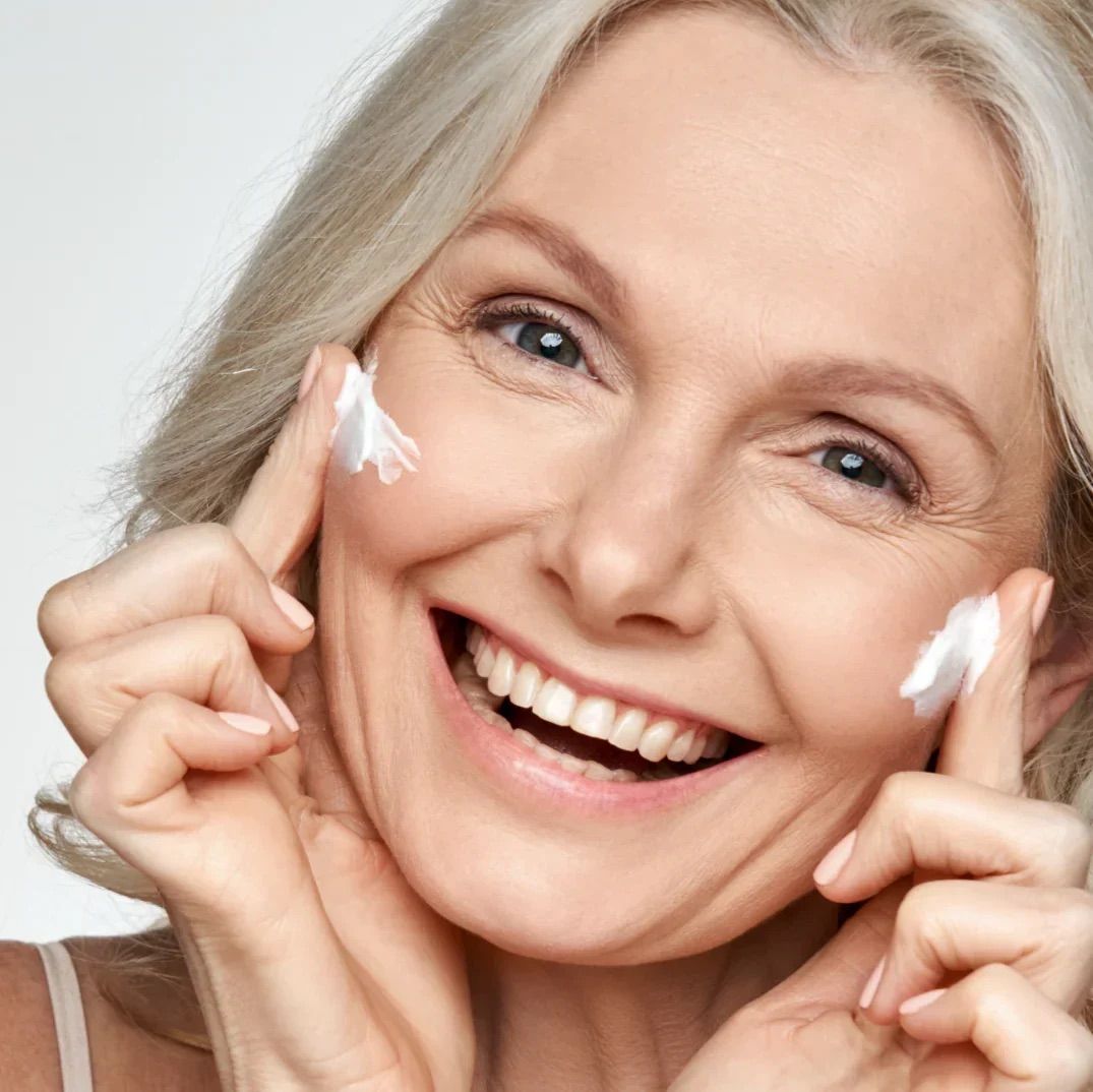 Ältere Frau mit Creme auf den Fingern. Sie lächelt. Creme auf Wangen aufgetragen.