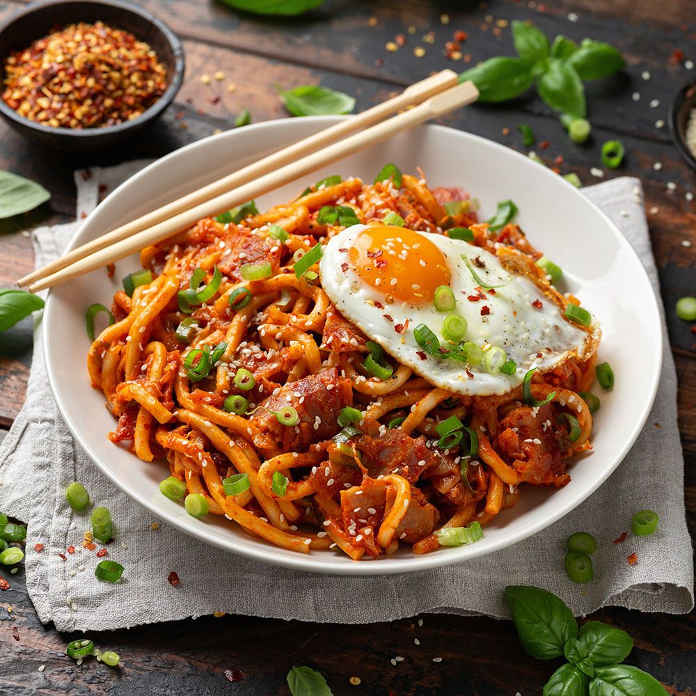 Nudeln mit Spiegelei, Kimchi und Gemüse in einer weißen Schüssel. Essstäbchen liegen daneben. Auf einem Holztisch.