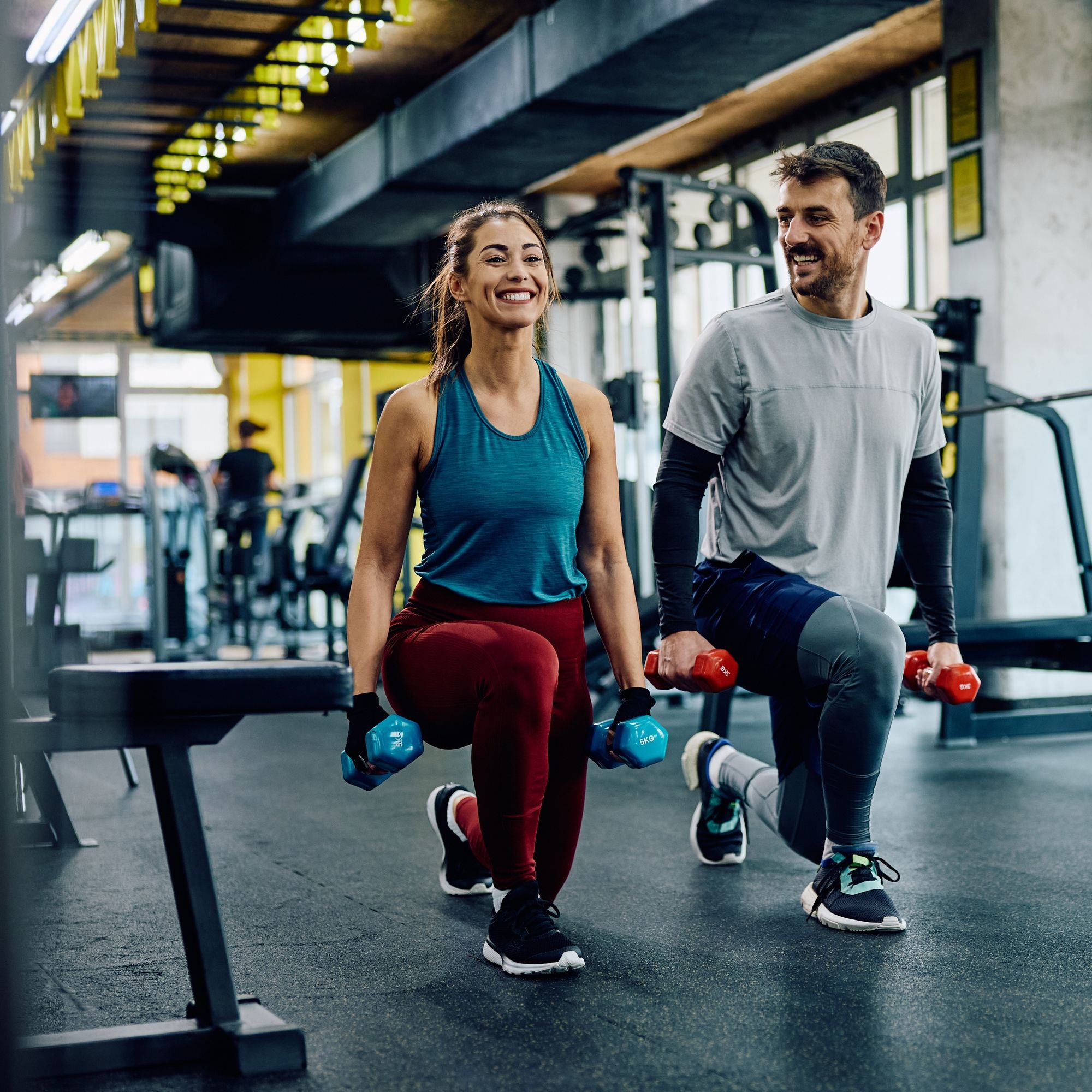 Zwei Personen trainieren im Fitnessstudio. Sie machen Ausfallschritte mit Kurzhanteln. Beide lächeln.