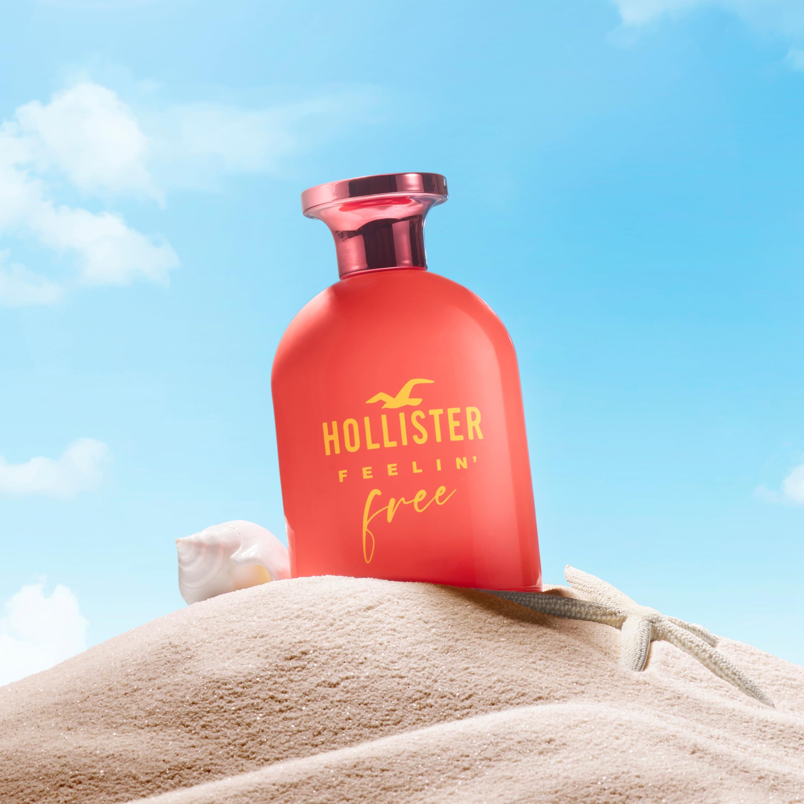 Roter Flakon mit gelbem Schriftzug. Aufschrift: Hollister Feelin' Free. Rosa Verschluss. Hintergrund: Sand.