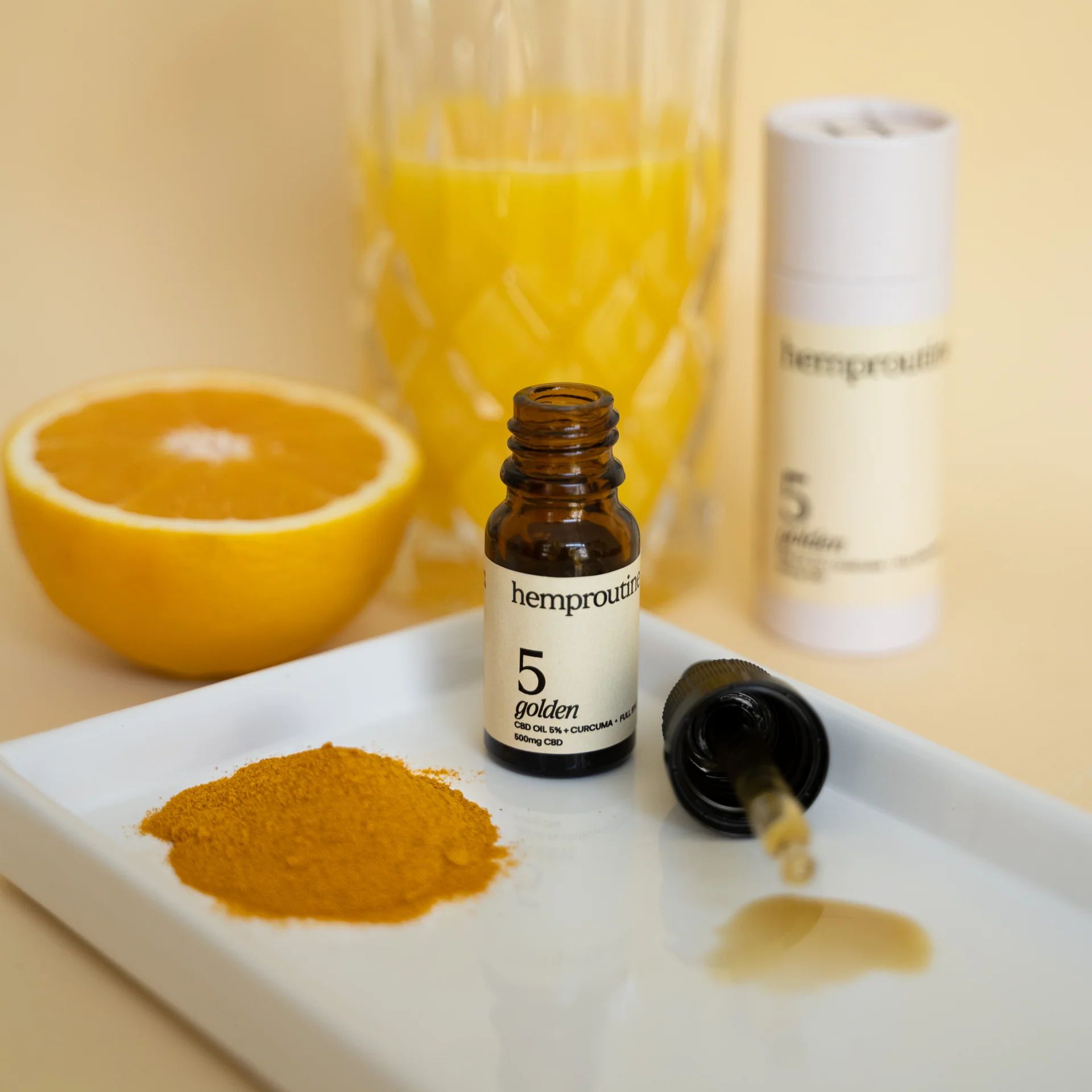 Braune Glasflasche mit Tropfer, daneben ein Glas Orangensaft, eine halbe Orange und Kurkumapulver auf weißem Tablett.