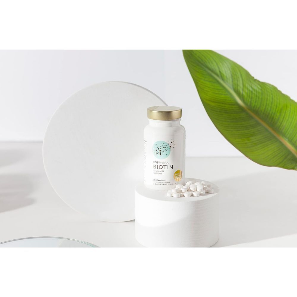 Weiße Flasche mit goldfarbenem Deckel. Aufschrift: Cosphera Biotin Vitamin B7 Tabletten. Daneben Tabletten auf einem Podest. Weißer Hintergrund.