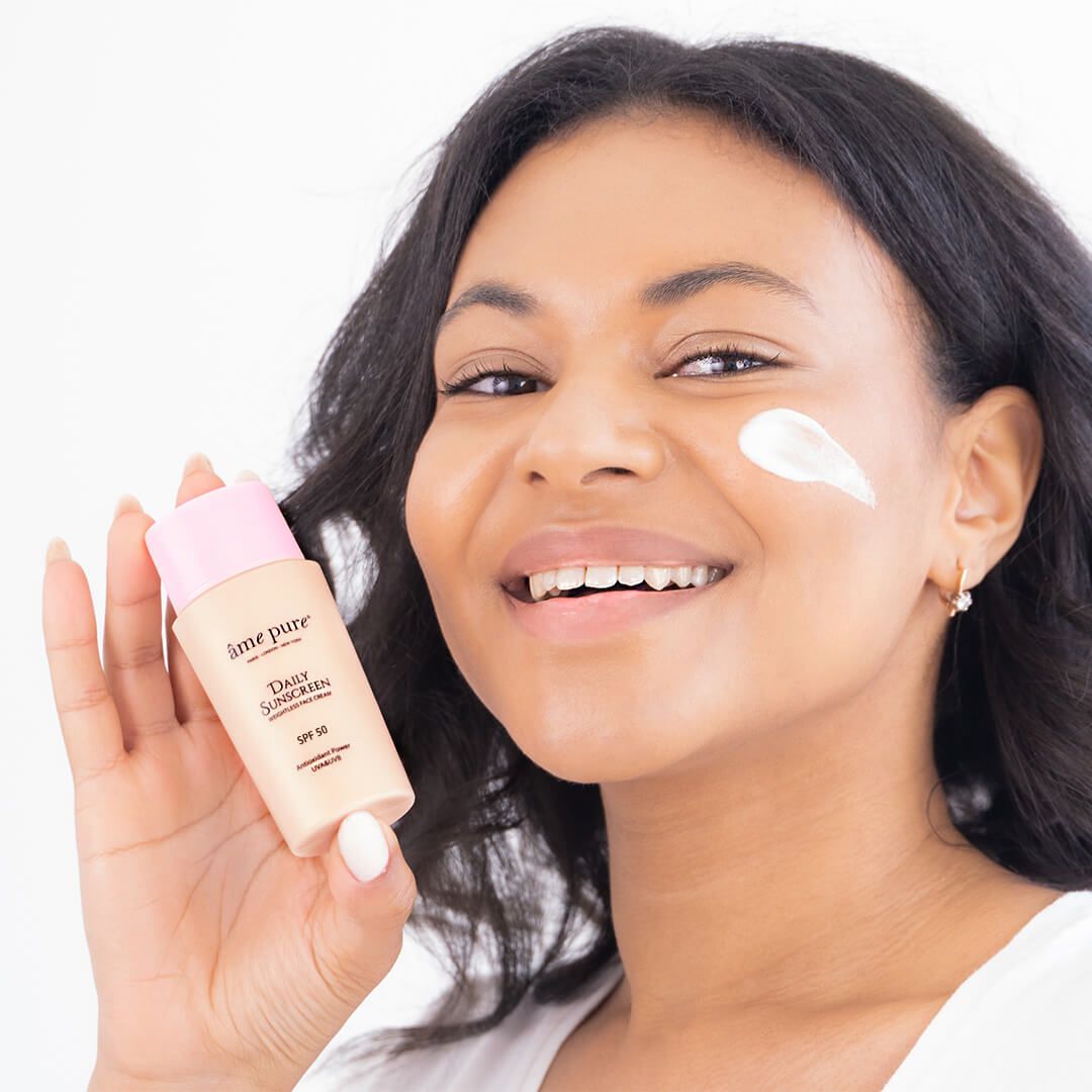 Frau mit Âme Pure Daily Sunscreen Flasche und Sonnencreme auf der Wange, SPF 50.
