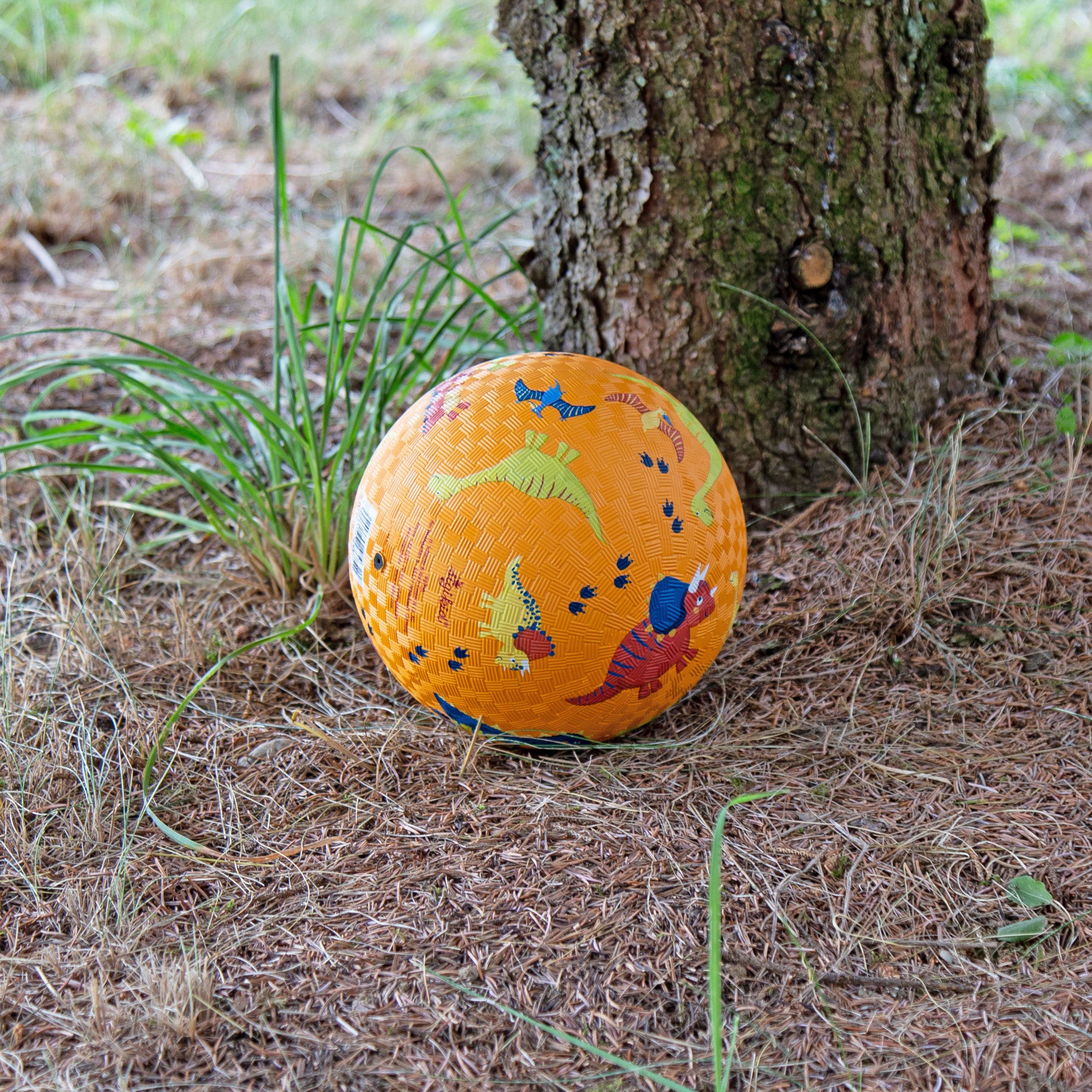 Gelber Ball mit Dinosaurier-Motiven liegt auf trockenem Gras unter einem Baum. Quadratisches Muster auf der Oberfläche.