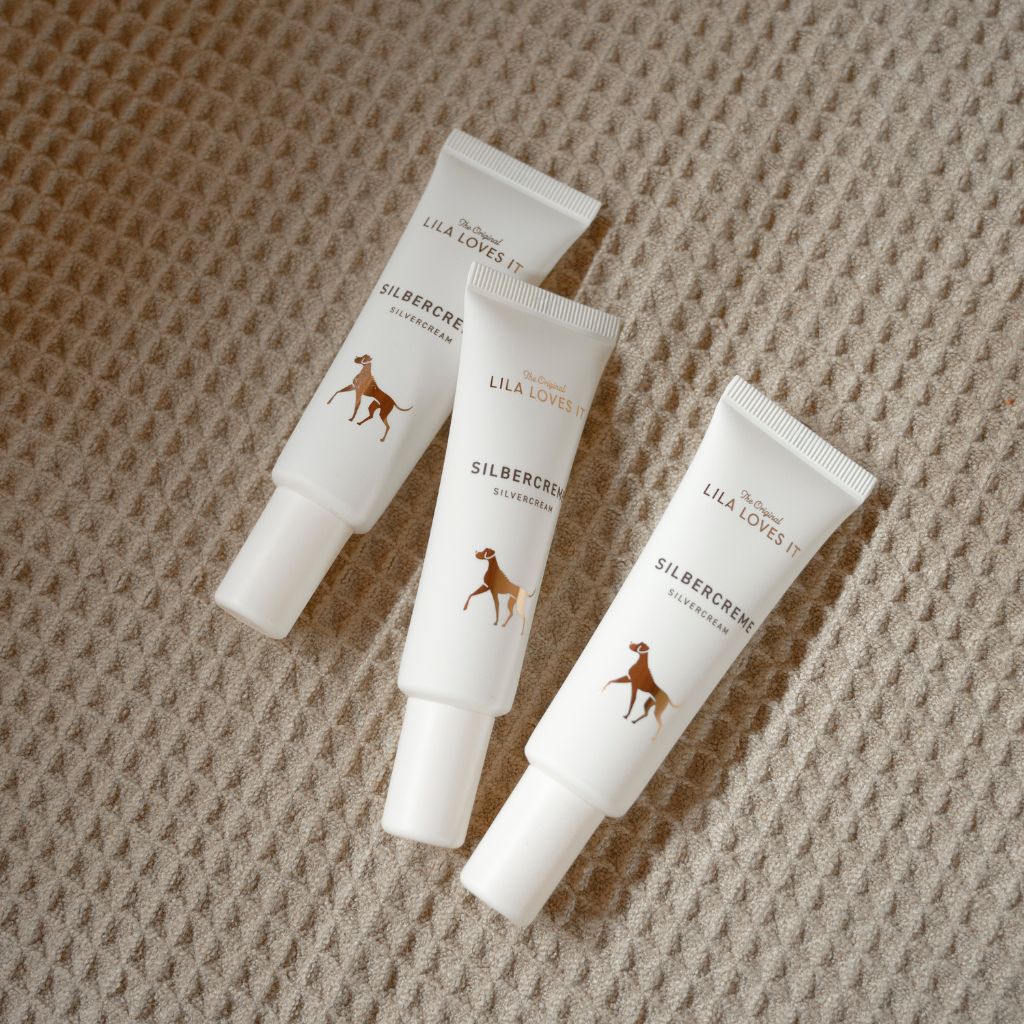 Drei Creme-Tuben "LILA LOVES IT" und "SILBERCREME". Abbildung eines Hundes. Auf hellem Untergrund.