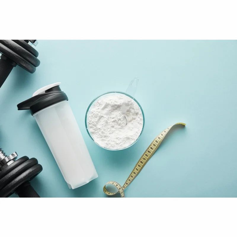 Shaker, Schüssel mit weißem Pulver, Messbecher und Maßband auf blauem Hintergrund. Fitness-Kontext.