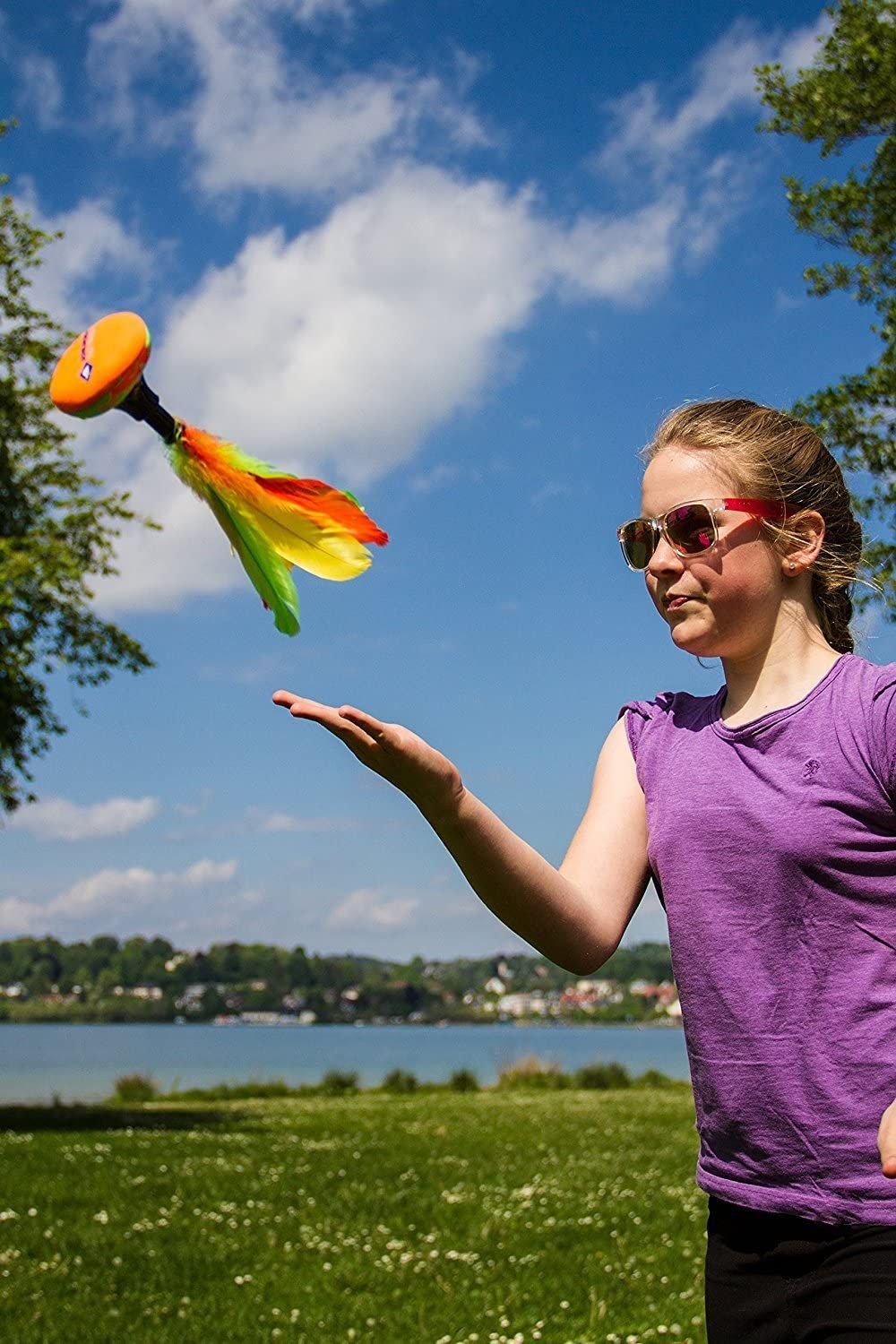 Mädchen wirft Peteca-Shuttle. Gelbe, grüne und orangefarbene Federn. Kind trägt Sonnenbrille und lila Oberteil.