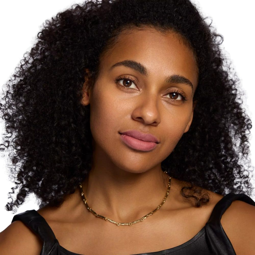 Porträt einer Frau mit rosafarbenem Lippenstift. Sie trägt eine goldene Halskette und ein schwarzes Oberteil.