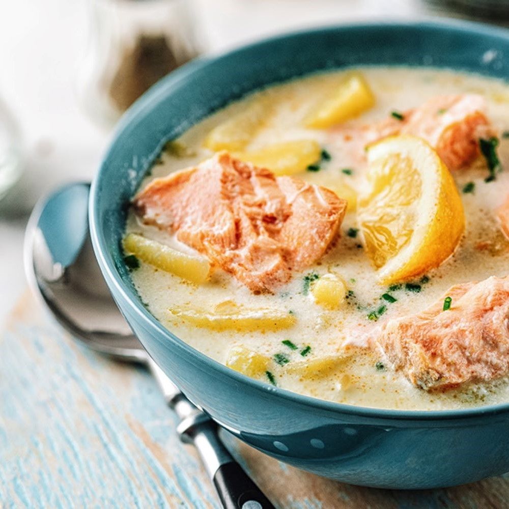 Blaue Schüssel mit Lachssuppe. Lachs, Kartoffeln und Zitronenscheibe. Löffel und Holzbrett.