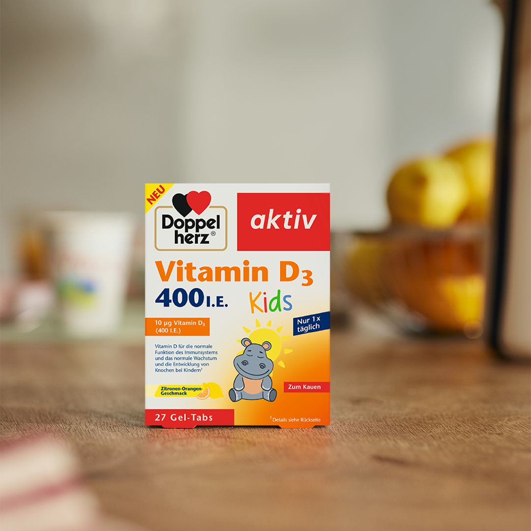 Doppelherz aktiv Vitamin D3 400 I.E. Kids Packung auf einem Holztisch, leicht unscharfer Hintergrund mit Küchenutensilien.