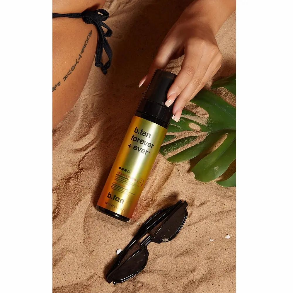 Selbstbräunungsmousse-Flasche mit schwarzem Pumpkopf auf Sand. Sonnenbrille und Blatt daneben. Teilweise sichtbare Haut und Tattoo.