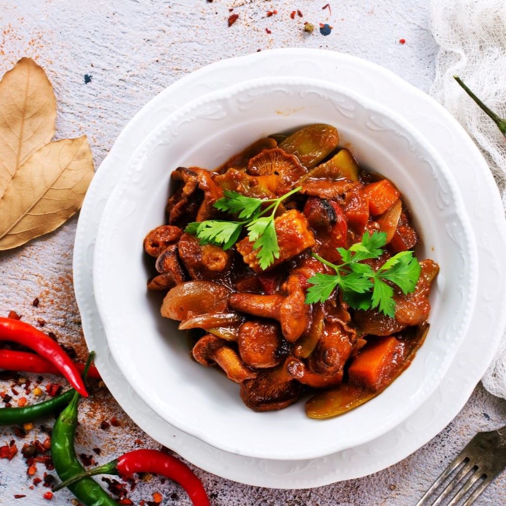 Gulasch in weißer Schüssel. Enthält Gemüse und Fleisch. Mit Petersilie garniert. Auf hellem Untergrund. Rote und grüne Chili.