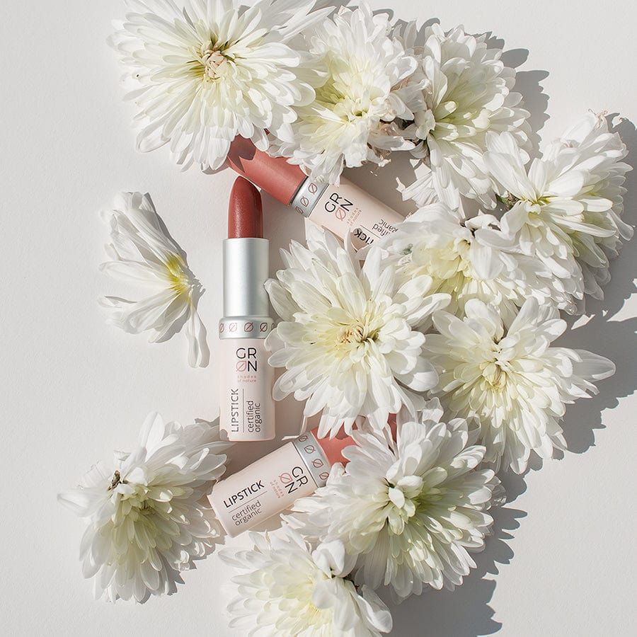 Drei Lippenstifte, umgeben von weißen Chrysanthemen. Zwei Lippenstifte mit Deckel, einer geöffnet. Aufschrift: GRN, Lipstick, certified organic.