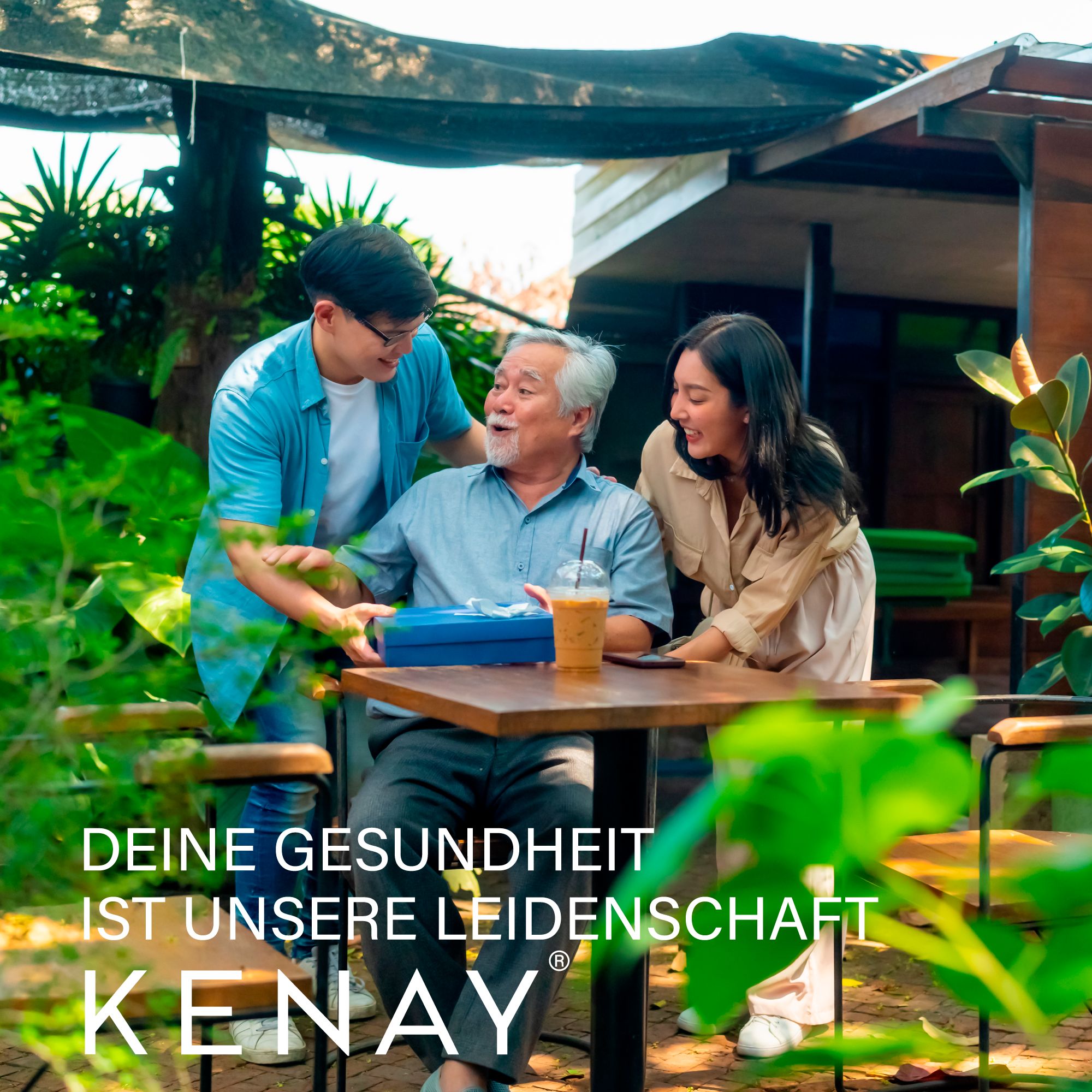 Drei Personen an einem Tisch. Eine Person überreicht eine Geschenkbox. Text: DEINE GESUNDHEIT IST UNSERE LEIDENSCHAFT. KENAY® Logo.
