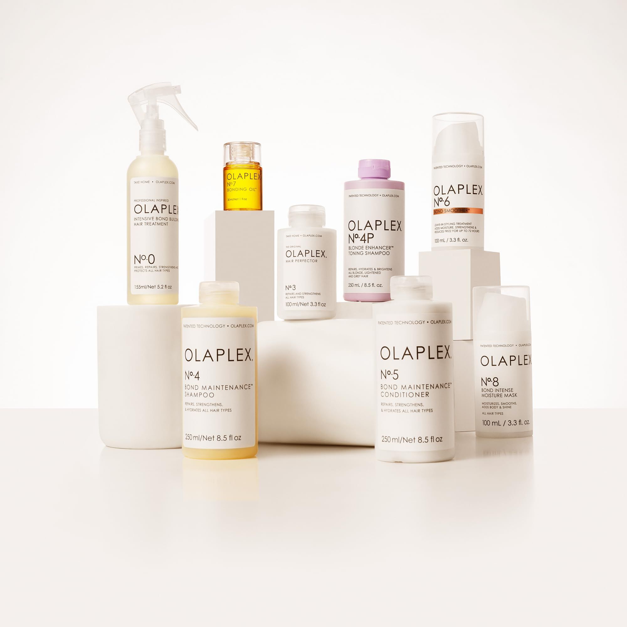 Verschiedene OLAPLEX-Haarpflegeprodukte auf weißen Podesten. Enthalten Flaschen, Tiegel und Sprays. Aufschrift: OLAPLEX.