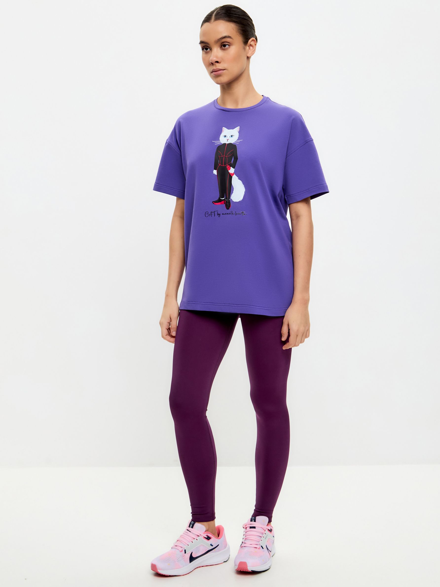 Dunkelviolette Leggings. Frau trägt lila T-Shirt mit Katzenmotiv. Pinke Schuhe. Stehende Pose.
