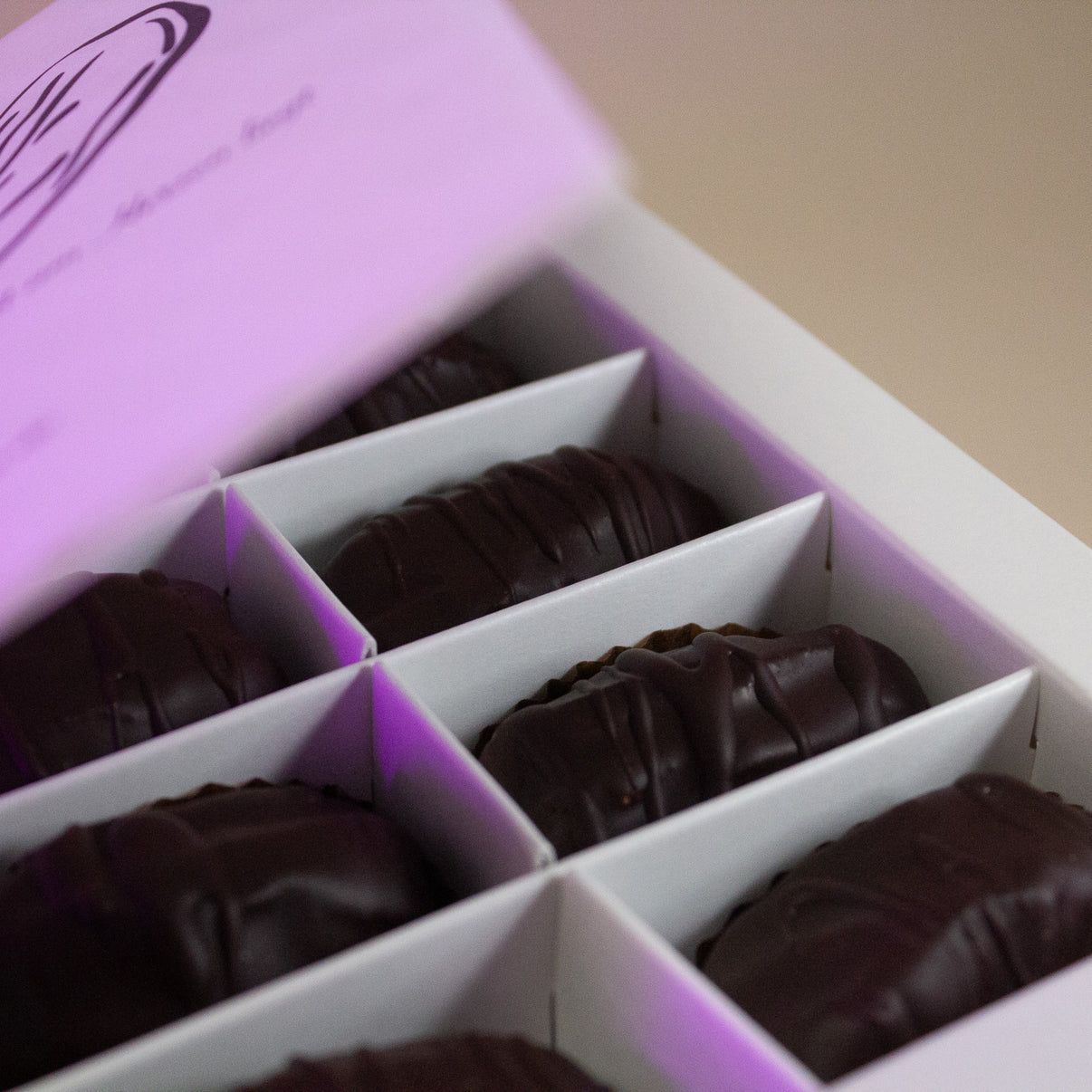 Nahaufnahme von sechs dunklen Schokoladen-Datteln in einer weißen Geschenkbox.
