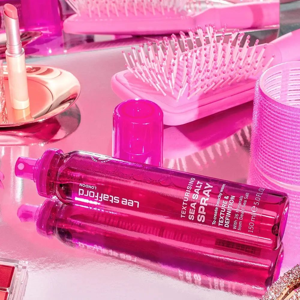 Fuchsiafarbene Sprühflasche Lee Stafford Texturising Sea Salt Spray auf glänzender Oberfläche, umgeben von Haarbürsten.