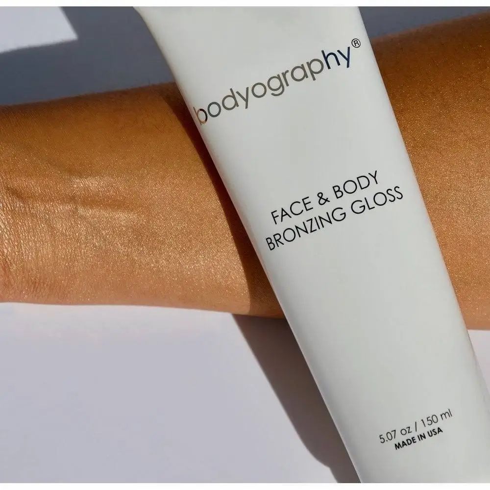 Arm mit gebräunter Haut neben einer Tube. Aufschrift: BODYOGRAPHY FACE & BODY BRONZING GLOSS. Volumenangabe: 5.07 oz / 150 ml.