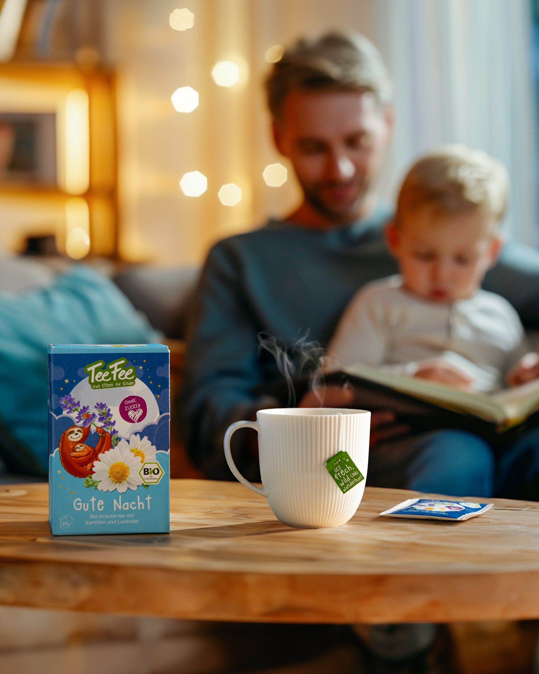 TeeFee-Verpackung auf Tisch. Tasse mit Teebeutel. Mann und Kind lesen im Hintergrund.