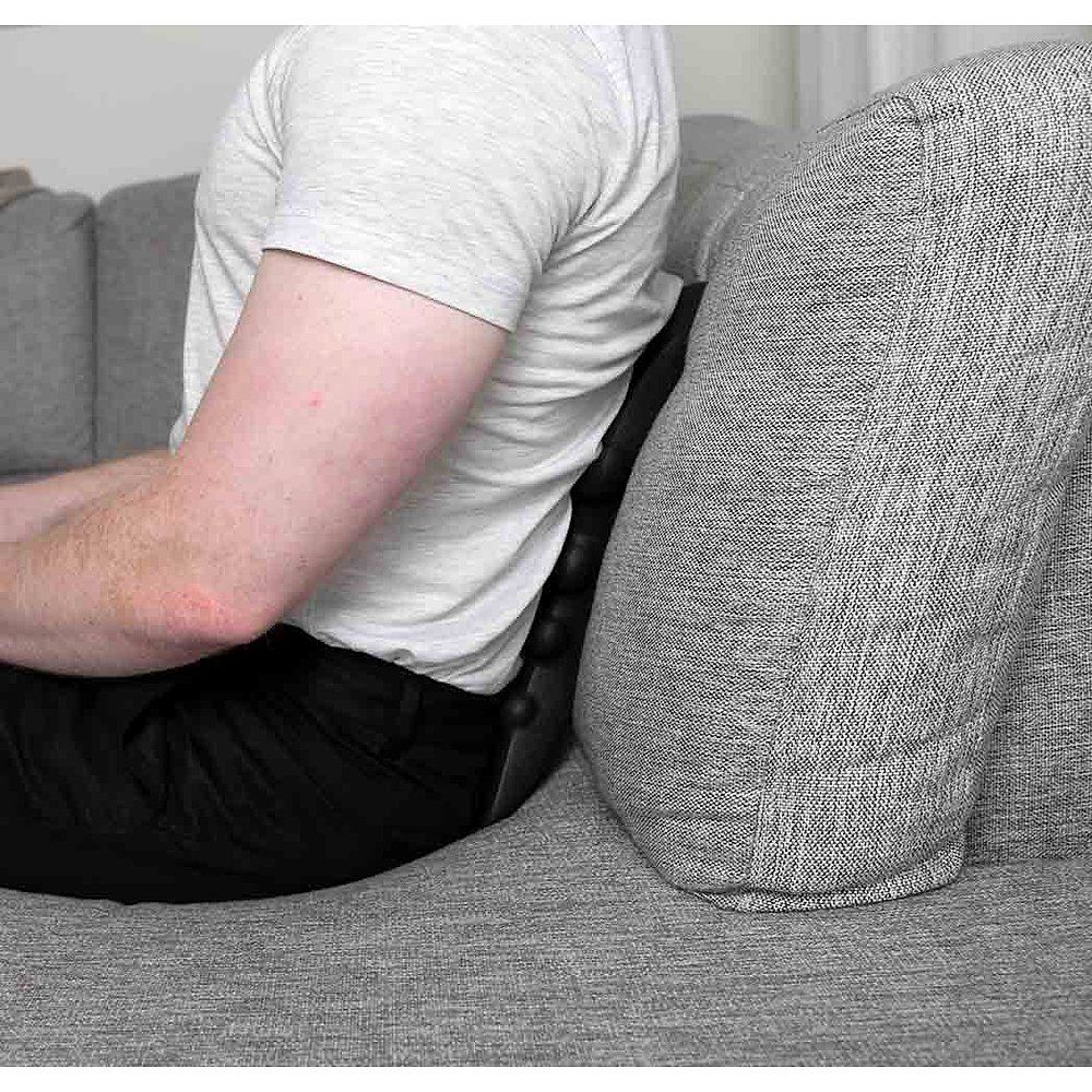 Person sitzt auf einem Sofa. Ein schwarzes Massagegerät ist zwischen Rücken und Sofa platziert. Das Gerät hat runde Noppen.