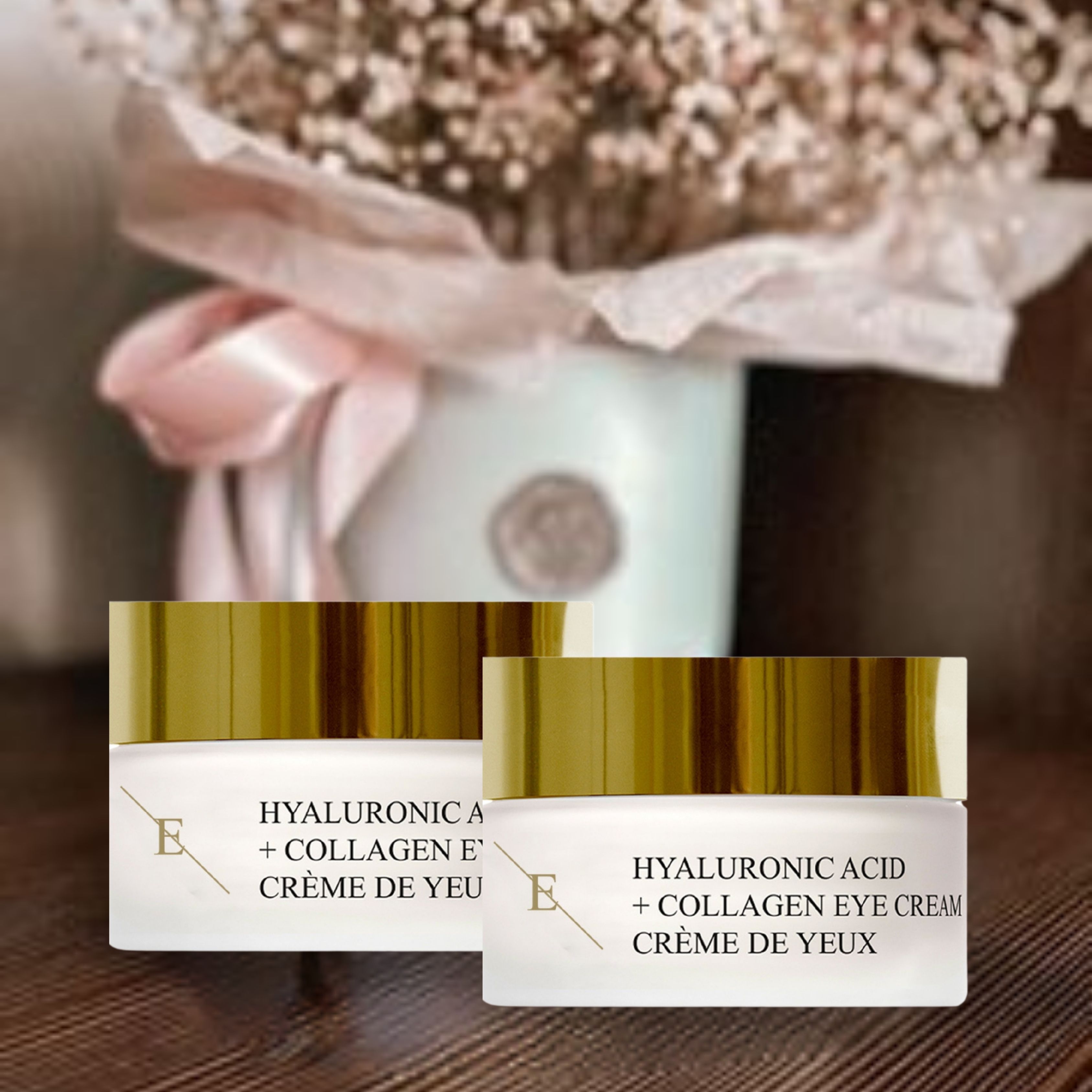 Zwei Tiegel Augencreme mit goldfarbenen Deckeln. Aufschrift: Hyaluronsäure + Kollagen Eye Cream. Creme de Yeux. Logo 'E'. Auf einem Tisch, mit Blumen im Hintergrund.