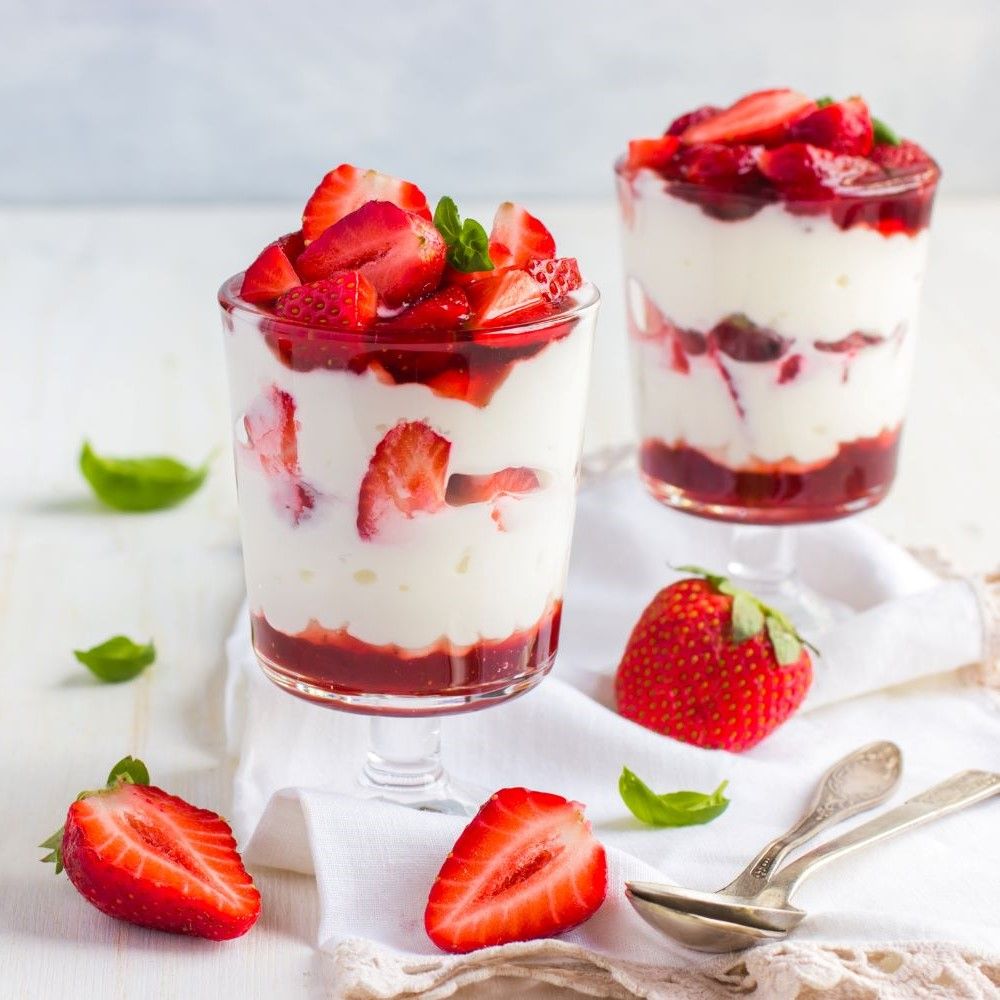 Zwei Gläser mit Erdbeeren und Sahne. Dekoriert mit Minze. Auf weißem Tuch. Daneben Löffel und halbe Erdbeere.