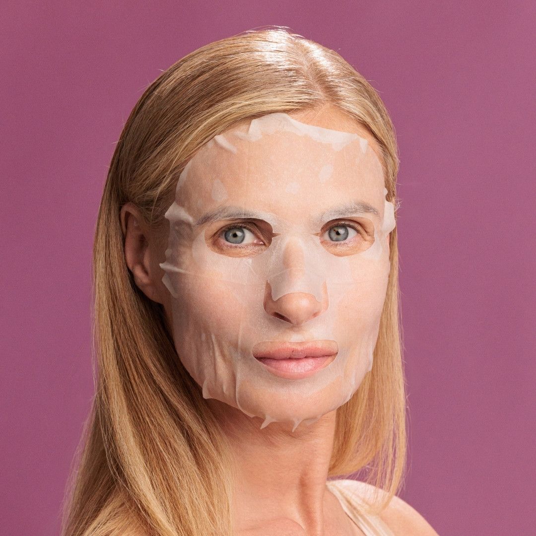 Frau mit Gesichtsmaske auf violettem Hintergrund. Maske bedeckt Gesicht mit Augen-, Nasen- und Mundöffnungen. Blondes Haar.
