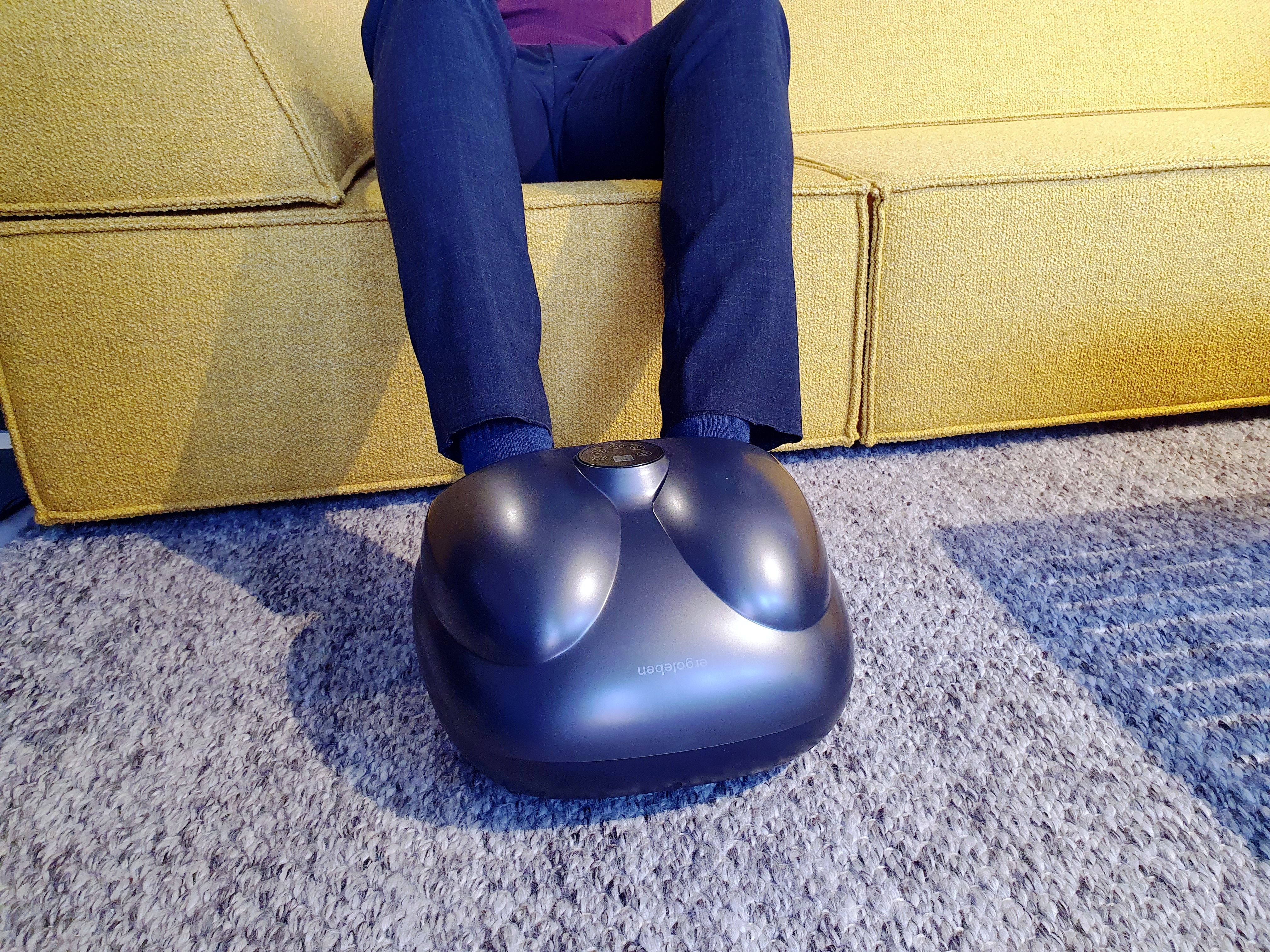 Vibrationsmassagegerät, universal, kabellos. Dunkelgraues Gerät, Füße auf Auflage. Auf Teppich, Person sitzt auf Sofa.