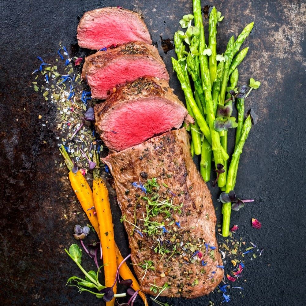 Gebratenes Rinderfilet, geschnitten. Mit Spargel, Karotten und Blüten garniert. Auf schwarzem Untergrund.