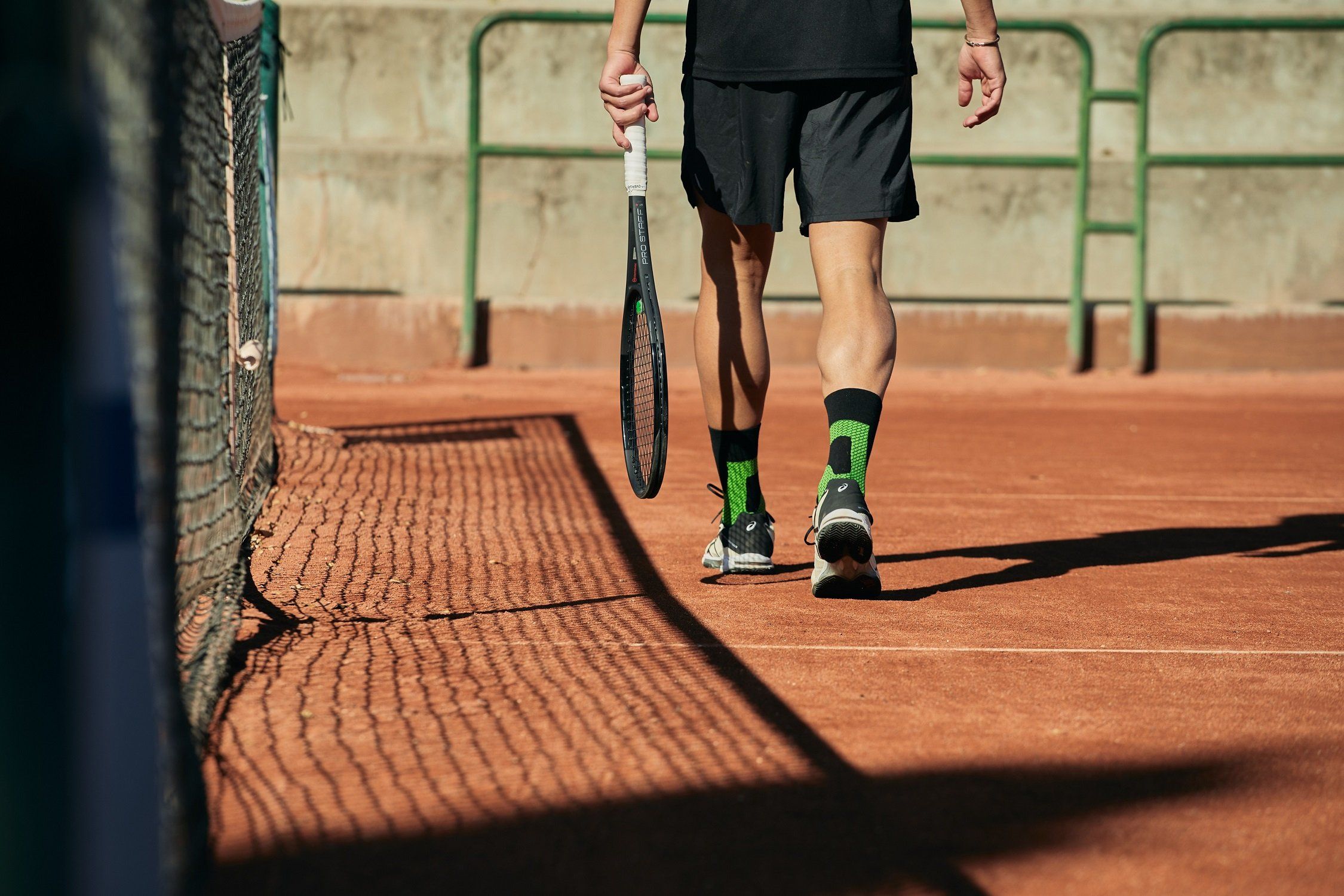 Una persona in piedi su un campo da tennis indossa un calzino stabilizzatore di caviglia Enforma. I calzini sono neri con dettagli verdi. La persona tiene in mano una racchetta da tennis.