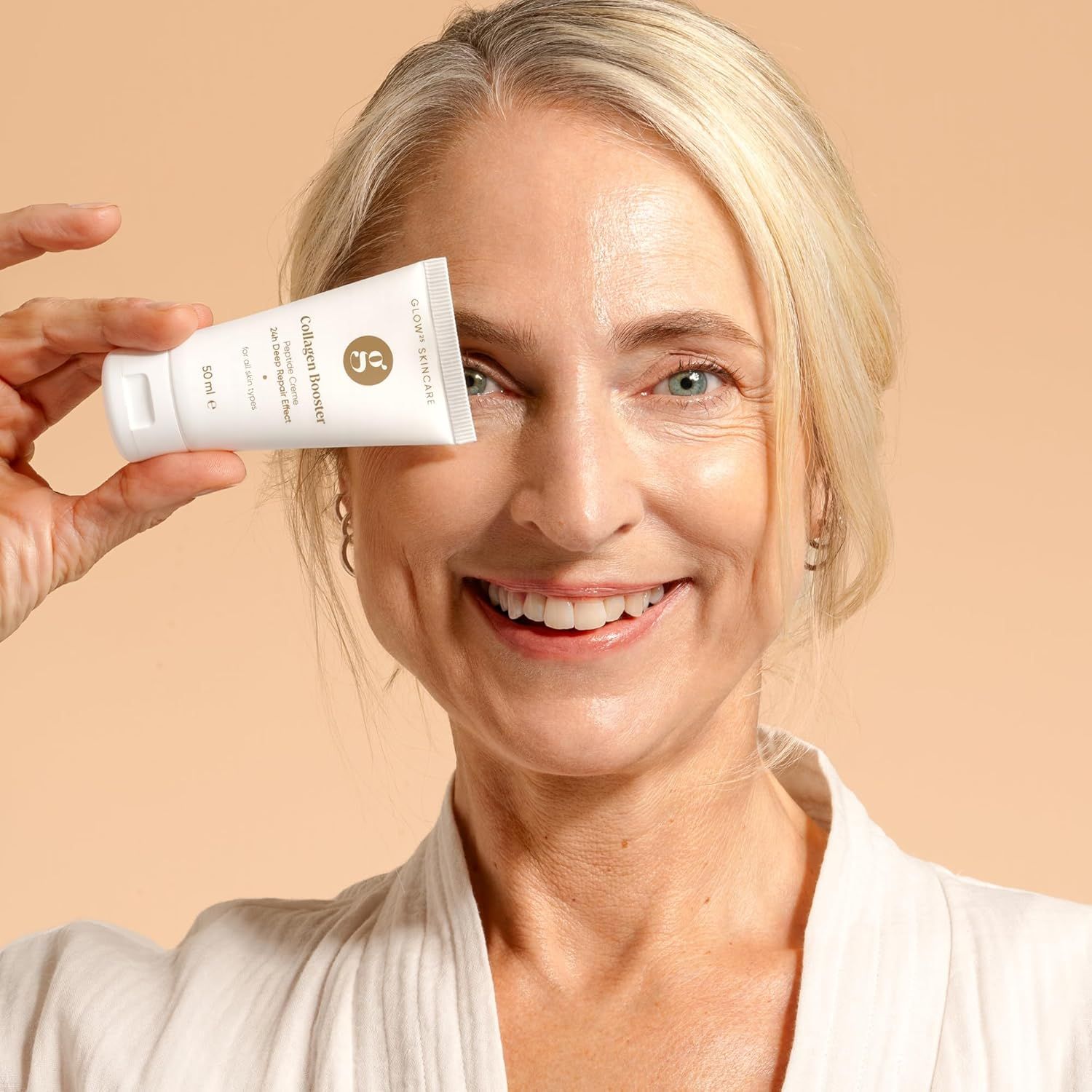 Une femme tient un tube blanc "Glow Skincare Collagen Booster" devant son œil. Sur le tube, on lit "50 ml". La femme sourit. Elle porte un peignoir blanc.