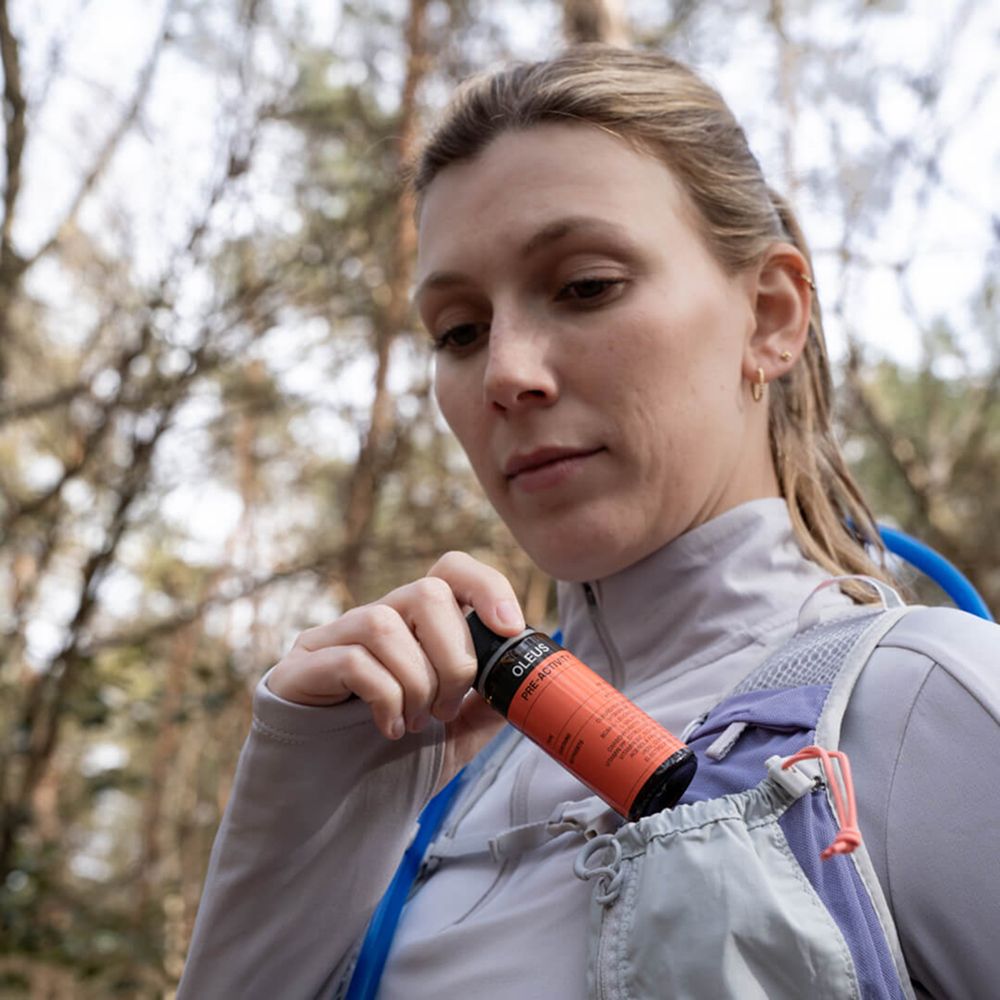 Frau mit Rucksack. Sie nimmt eine Flasche OLEUS aus einer Tasche. Wald im Hintergrund.