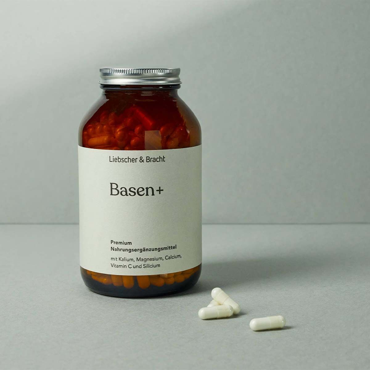 Braune Glasflasche mit silbernem Deckel und Etikett "Basen+". Daneben liegen drei weiße Kapseln.