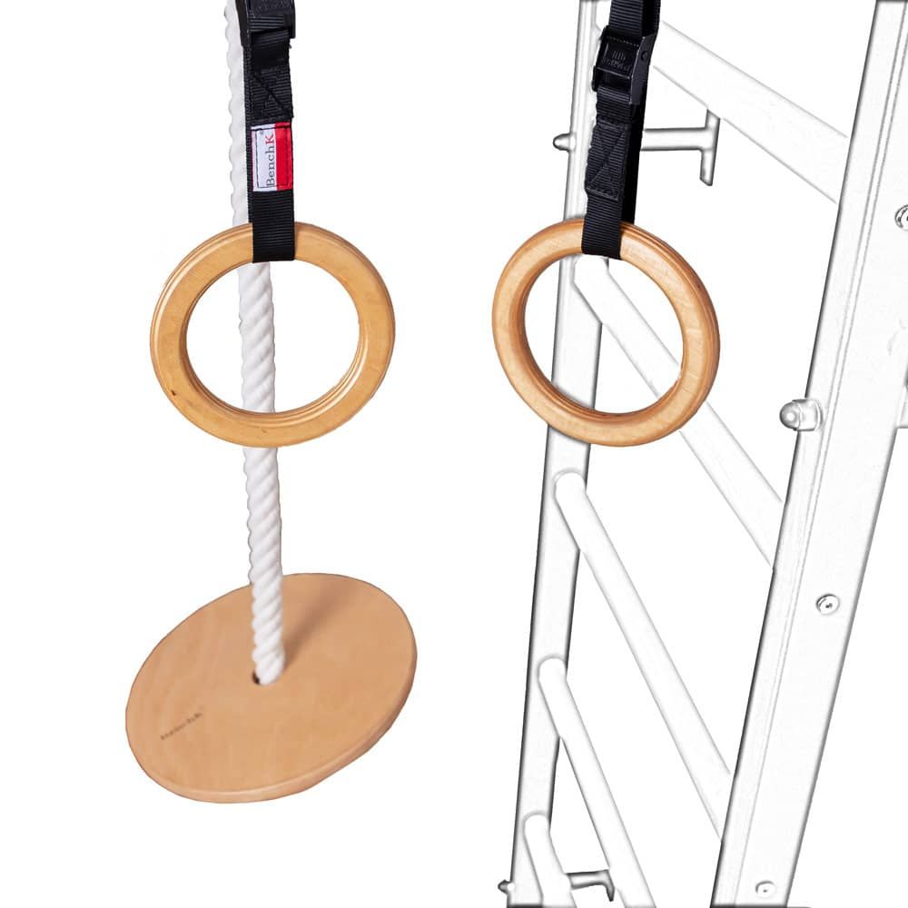 Holzringe mit schwarzen Gurten und weißem Seil. Ein Holzbrett ist am Seil befestigt. Der Hintergrund zeigt eine Sprossenwand.