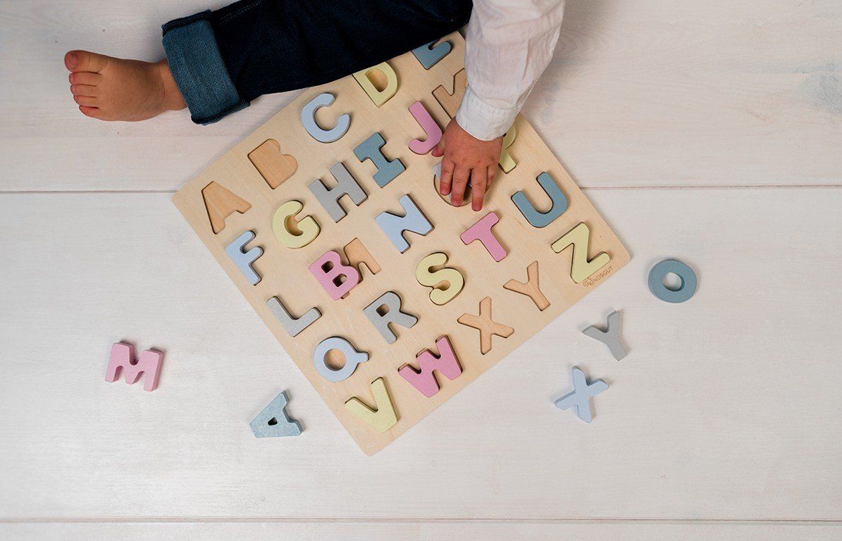 Kind sitzt neben Holzpuzzle mit Buchstaben. Einige Buchstaben sind herausgenommen. Helle Holzoberfläche.