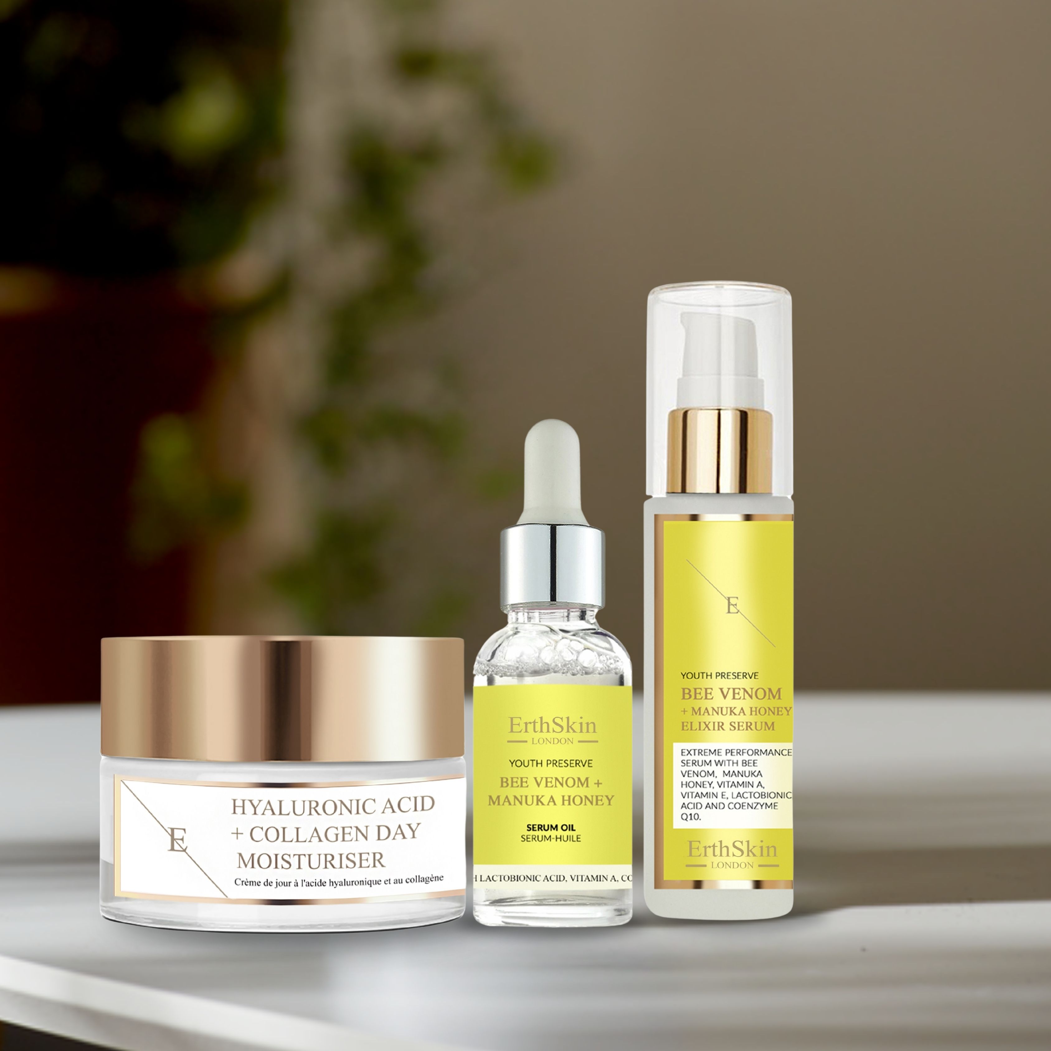 Drei Kosmetikprodukte: Tagescreme, Serumöl und Elixier-Serum. Aufschrift: Hyaluronsäure + Kollagen, Bee Venom + Manuka Honey. Auf Tisch.