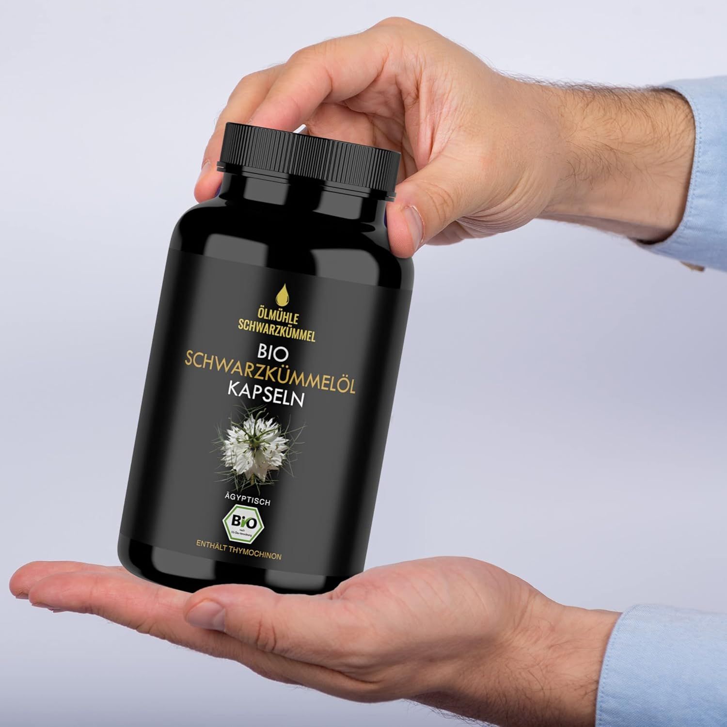 Hände halten eine schwarze Flasche mit BIO Schwarzkümmelöl Kapseln. Aufschrift: BIO Schwarzkümmelöl Kapseln. BIO-Siegel.