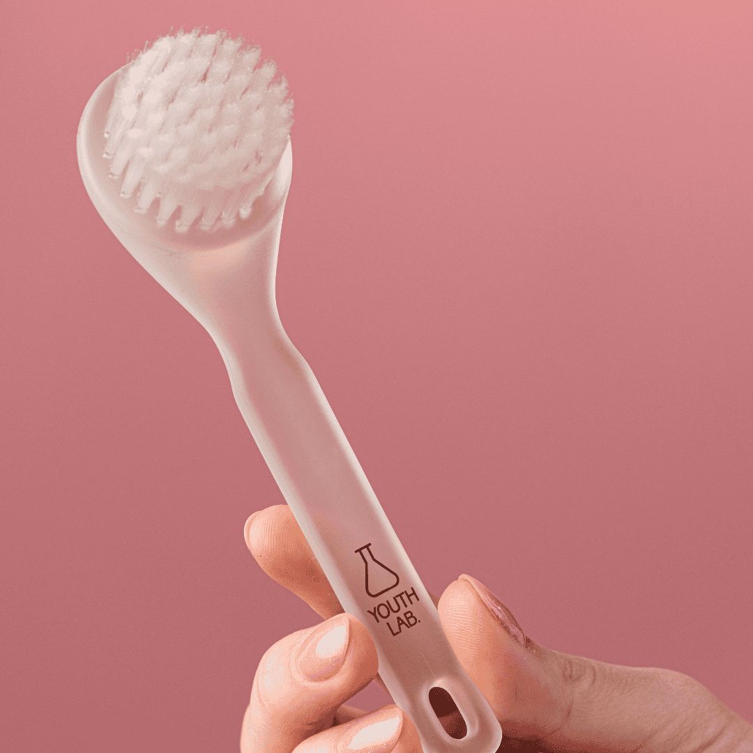 Heller, ovaler Reinigungsbürstenkopf mit Griff. YOUTH LAB Logo auf dem Griff. Hand hält das Werkzeug vor rosa Hintergrund.