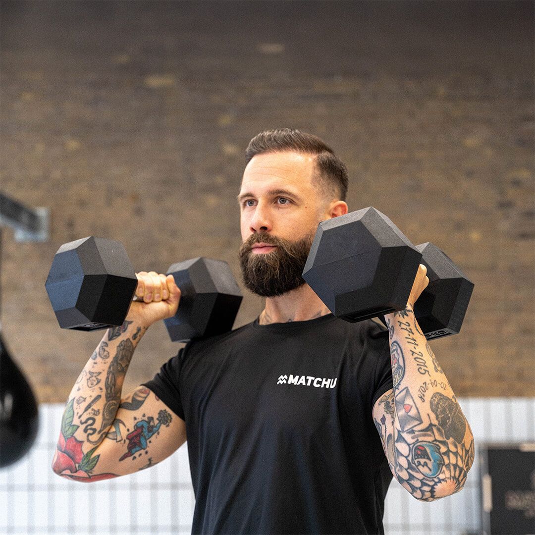 Mann hebt Kurzhanteln. Er trägt ein schwarzes Shirt mit dem Logo "MATCHU".