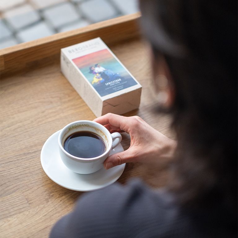 Person hält eine Tasse Kaffee und eine Packung BERGBRAND Spritztour. Holztisch.