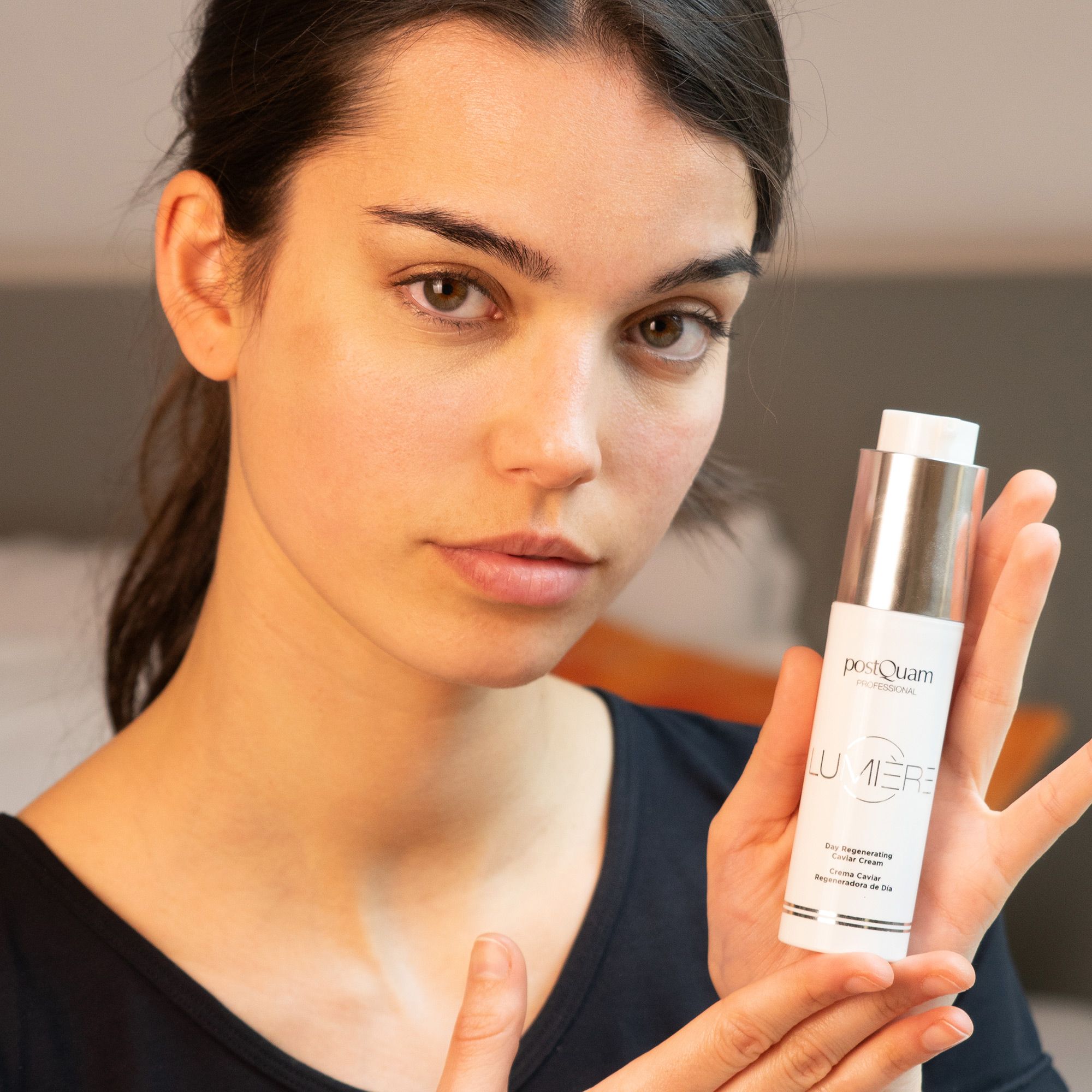 Frau hält Creme-Flakon. Aufschrift: PostQuam Professional, Lumière. Sie schaut in die Kamera.