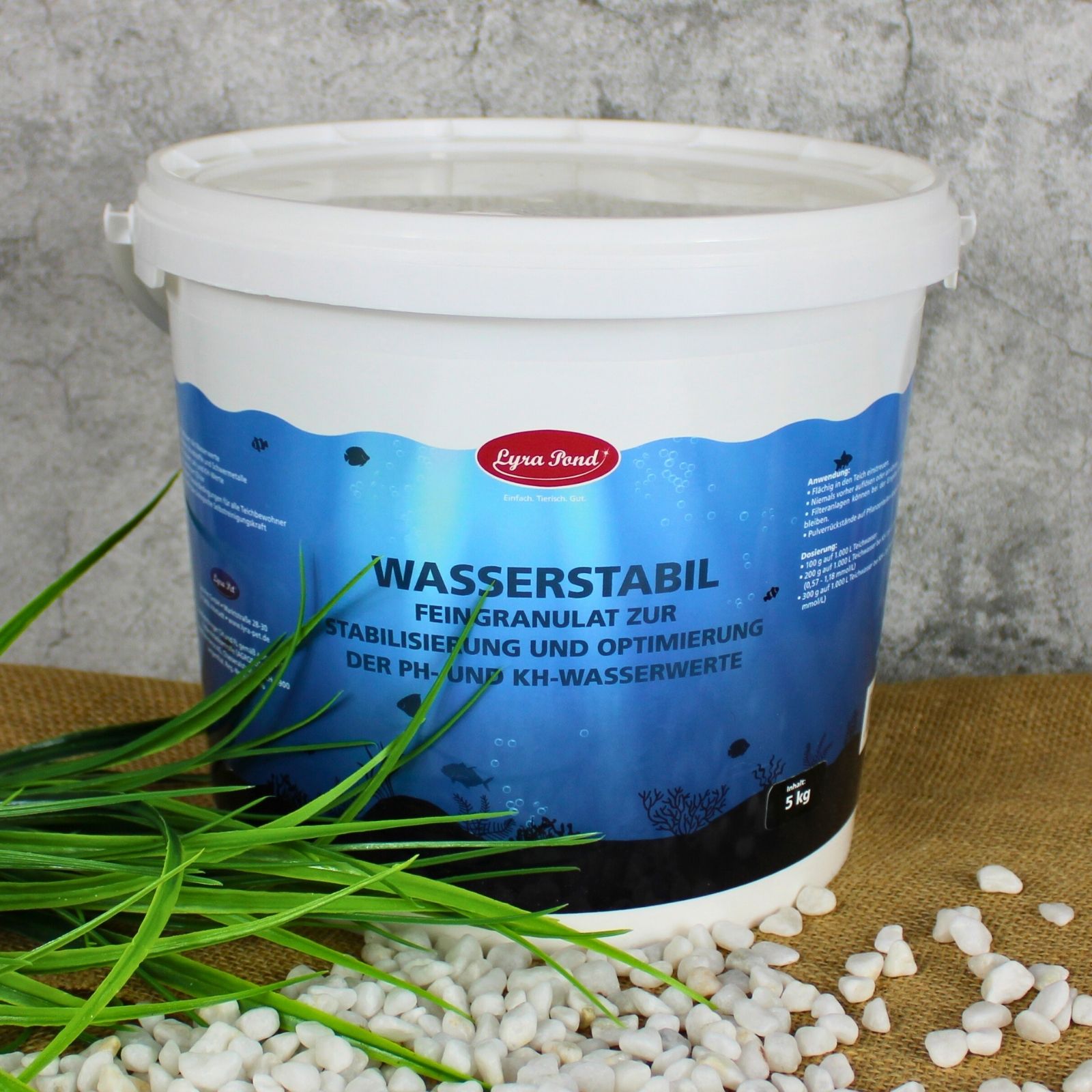 Weißer Eimer mit blauem Etikett 'Lyra Pond Wasserstabil Feingranulat'. Steht auf Jute mit grünen Grashalmen und weißen Kieselsteinen.