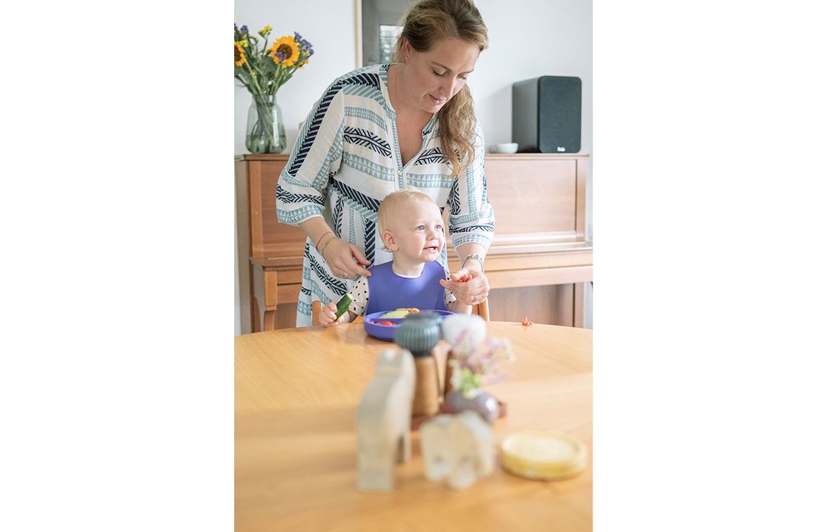 Baby mit lila Lätzchen am Tisch. Person befestigt Lätzchen. Auf dem Tisch Teller mit Essen.